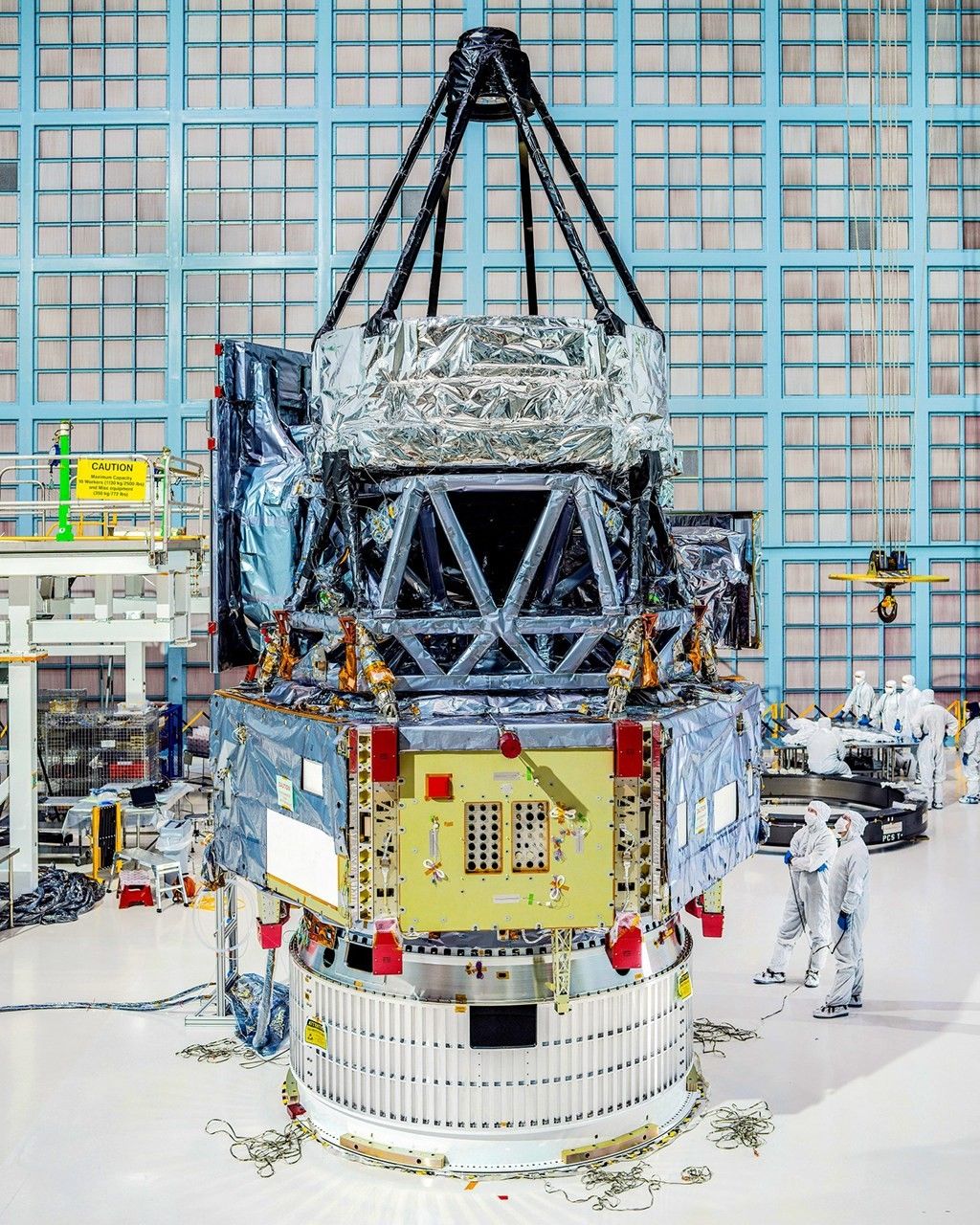 A large complex structure in a work room towers above workers clad in protective suits. At the top of the structure, six black struts extend to hold a small round mirror.