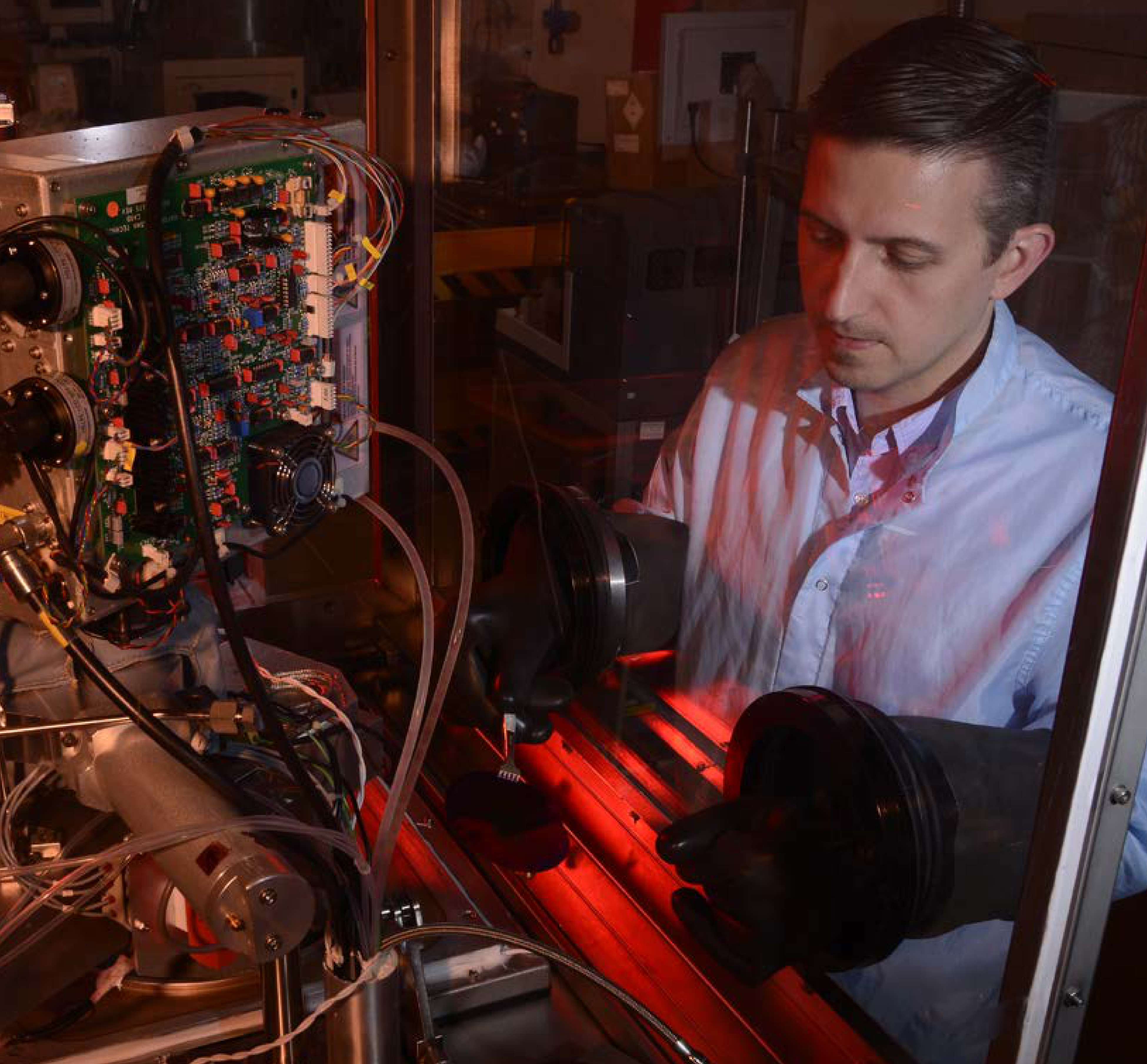 A person in a blue lab coat is working at a glovebox station that houses a large metal structure with numerous wires and connections.