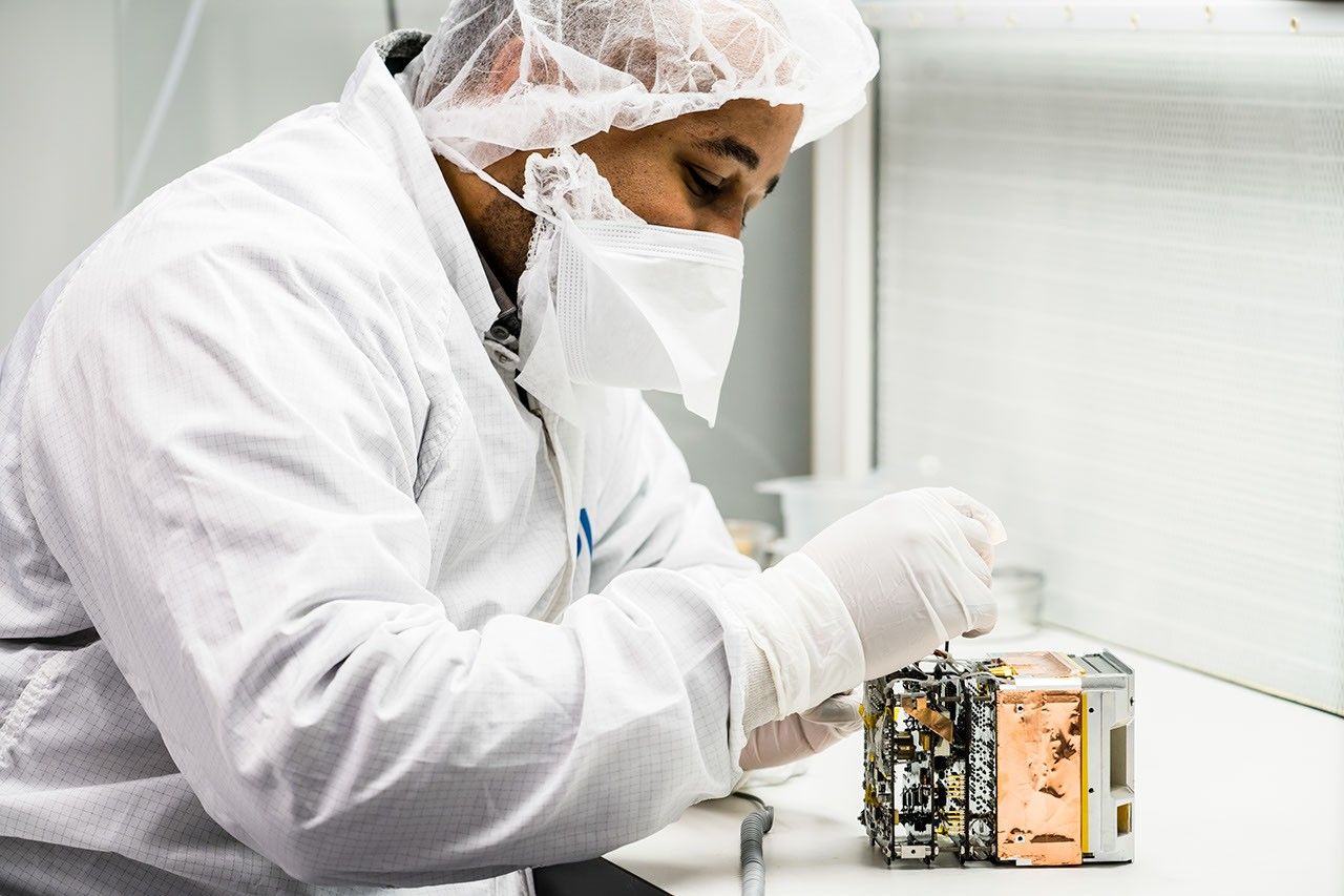 A man dressed in a white lab coat, hairnet, face mask, and gloves adjusts a fastener on a small instrument.