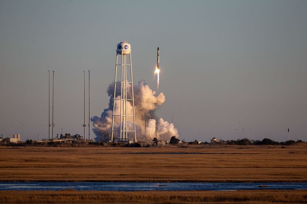 A Rocket Lab Electron rocket launching from a launch pad with a plume of smoke underneath and a water tower to the left. A marshland is in the foreground