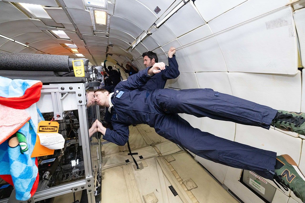 A person is floating in zero gravity horizontally inside a plane while looking inside a plexiglass container.