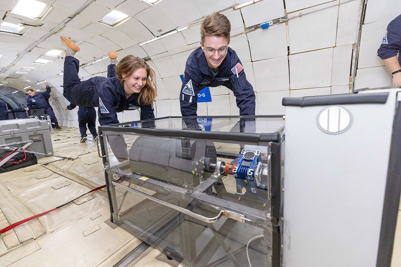 Two people are floating the zero gravity hovering above a clear case with an experiment inside.