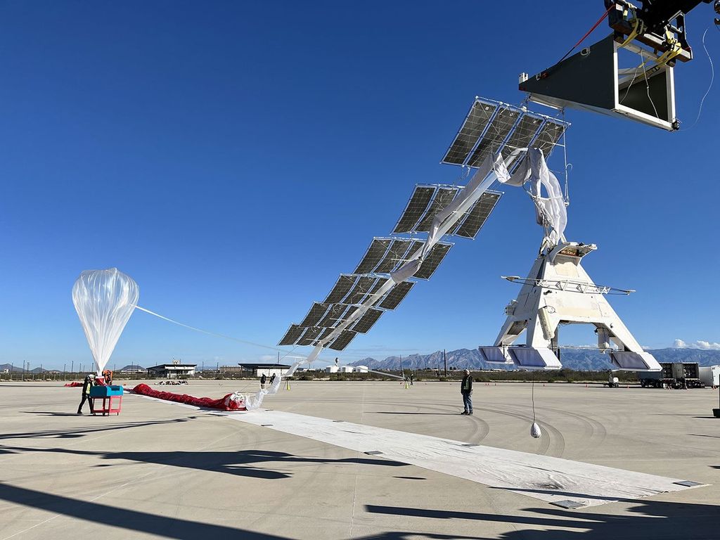 A partially inflated scientific balloon in the background with a payload made of many solar panels in the foreground.