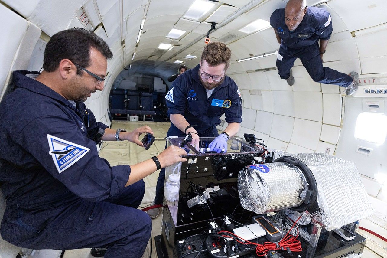 Three people working in zero gravity inside a plane around a box made of plexiglass.