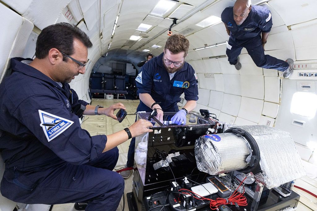 Three people working in zero gravity inside a plane around a box made of plexiglass.