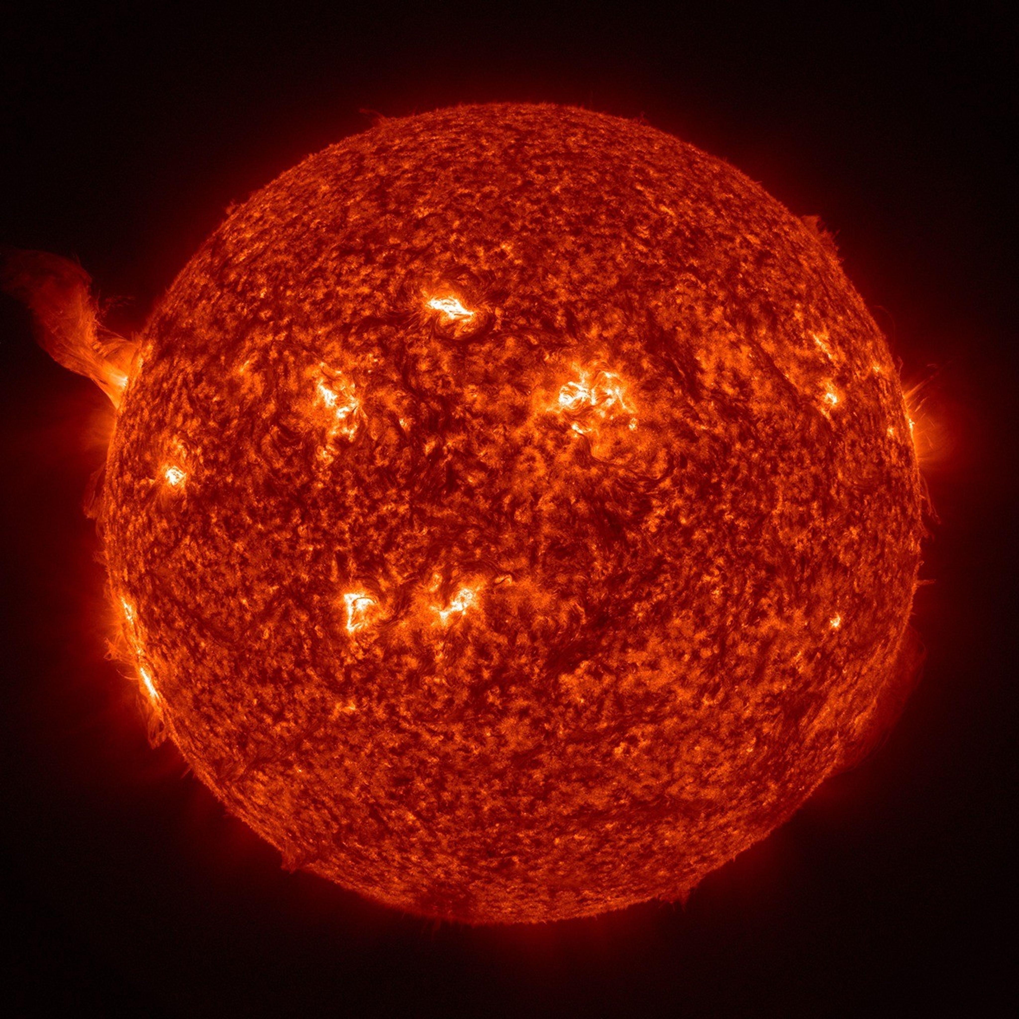 High-resolution image of the Sun captured in extreme ultraviolet light, showing a detailed view of the solar surface and atmosphere. Bright, active regions indicate intense magnetic activity, including sunspots, solar flares, and prominences. A large solar prominence is visible on the left edge, arching out into space.