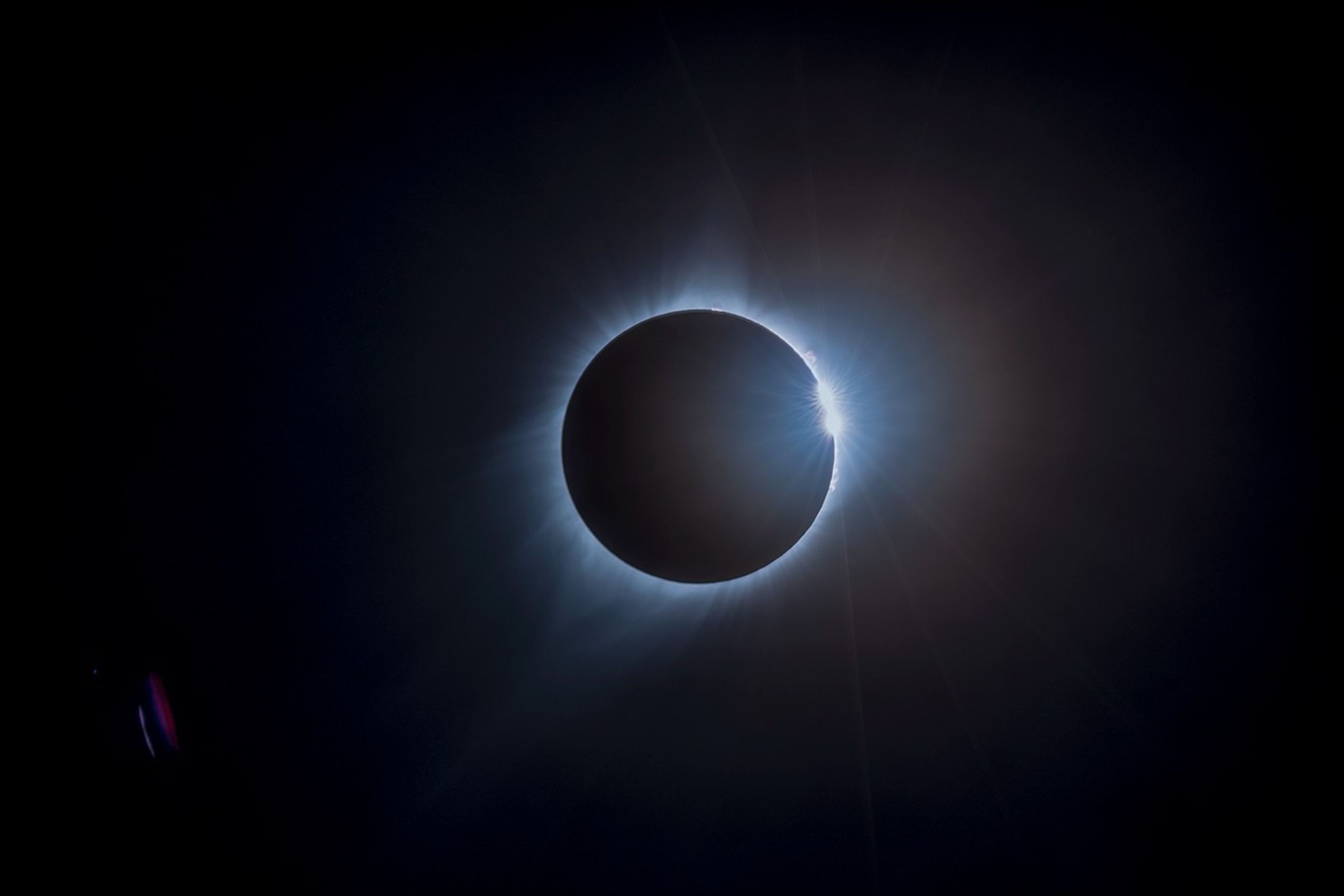 Set against a black background, a radiant gray ring of light appears around the black disk of the Moon during a total solar eclipse. On the right side of the ring, a small, bright, burst of light appears like a diamond.