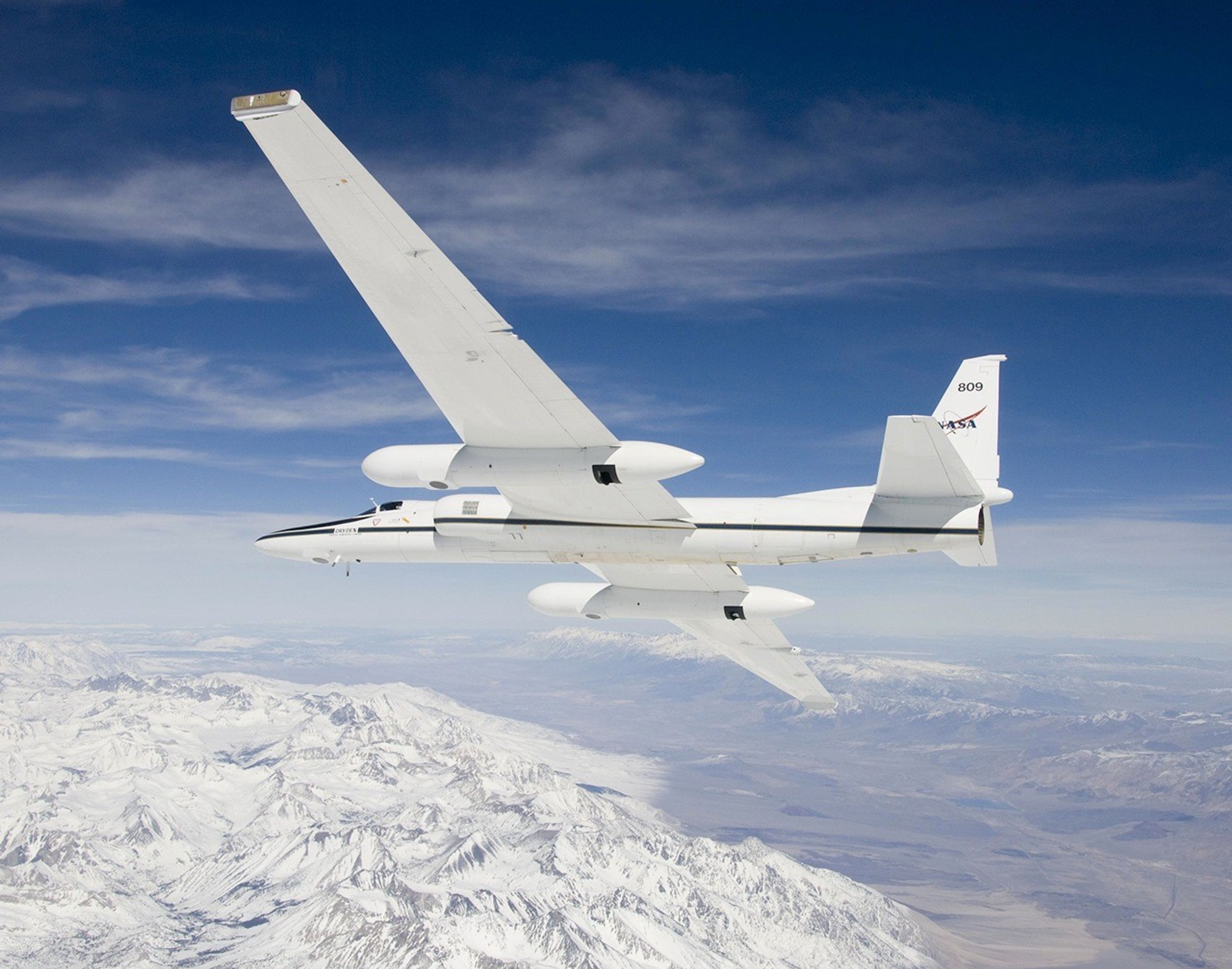 Against a bright blue sky streaked with clouds, a white airplane soars over snowy mountains.