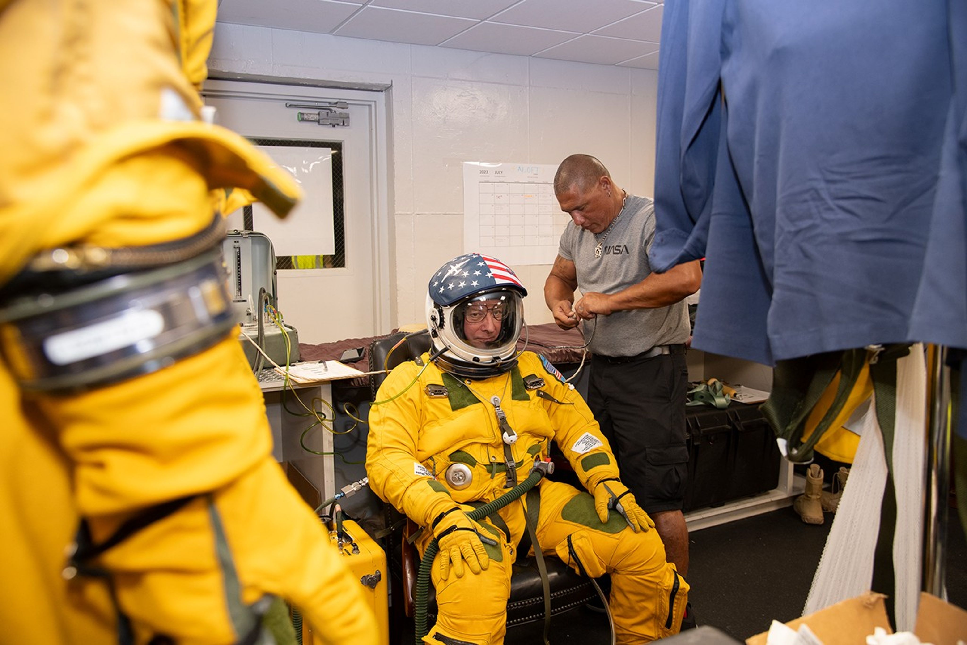 A man in a yellow spacesuit and American flag-helmet sits on a chair as another man in a NASA shirt connects tubing between the suit and equipment on a table.