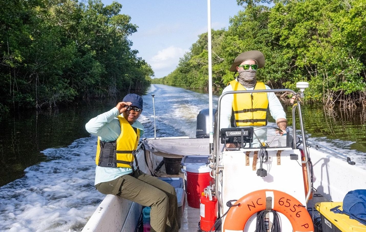 David Lagomasino drives a small, white skiff through a narrow channel lined with mangroves, which stick out of the water on either side of the boat as. David’s PhD student, Lola Babanawo, sits on the edge of the boat holding her cap.