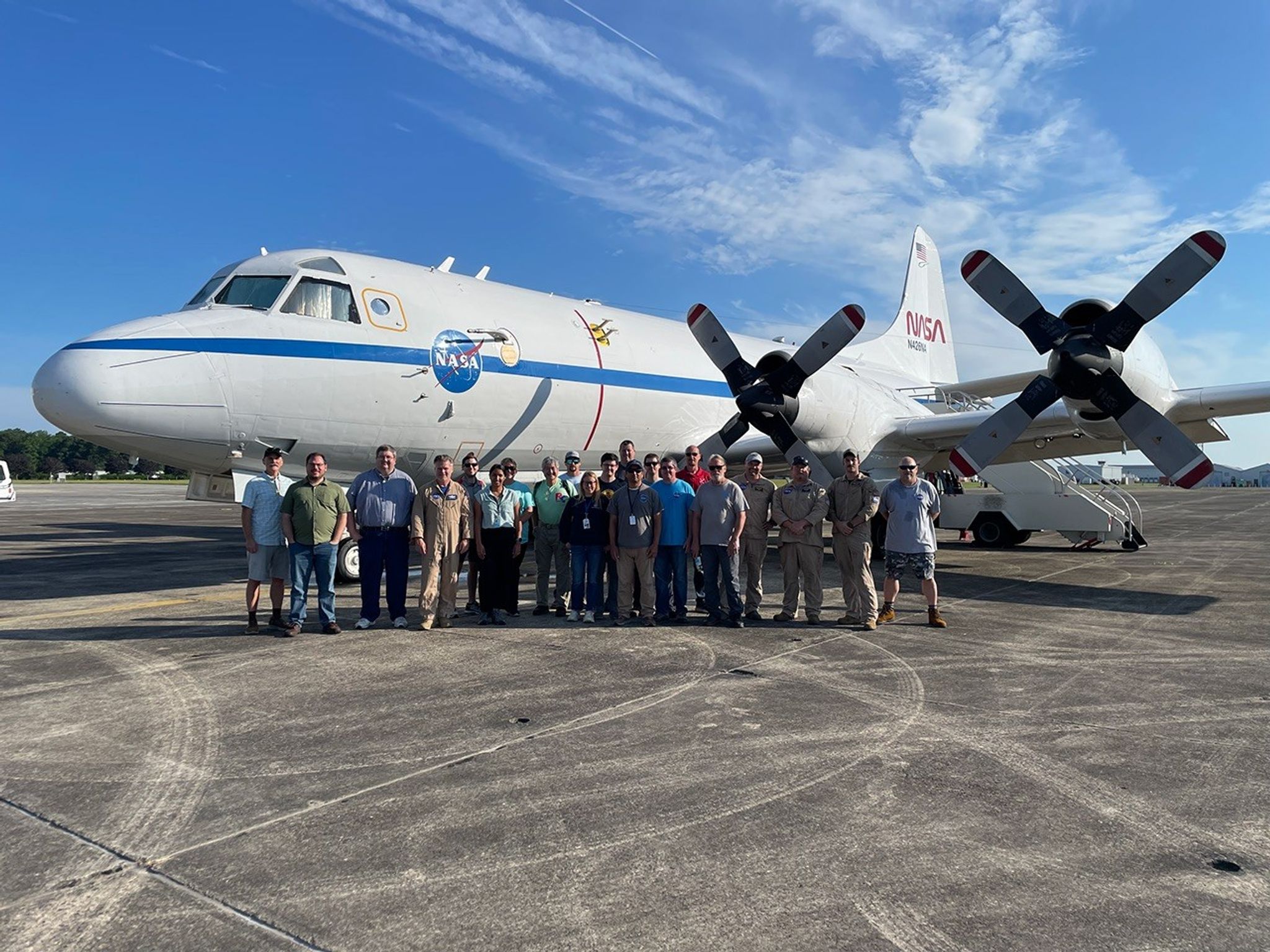 A group of twenty one people stand in front of a large, white aircraft with propellors. The NASA meatball logo is seen on the side of the aircraft. The people are all smiling and looking at the camera.