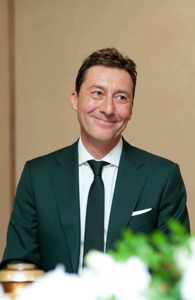 A man is centered in the image, looking slightly to the right of the camera and smiling. He has short, dark hair, and is wearing a dark green suit and matching tie. He is seen from the mid-torso up.