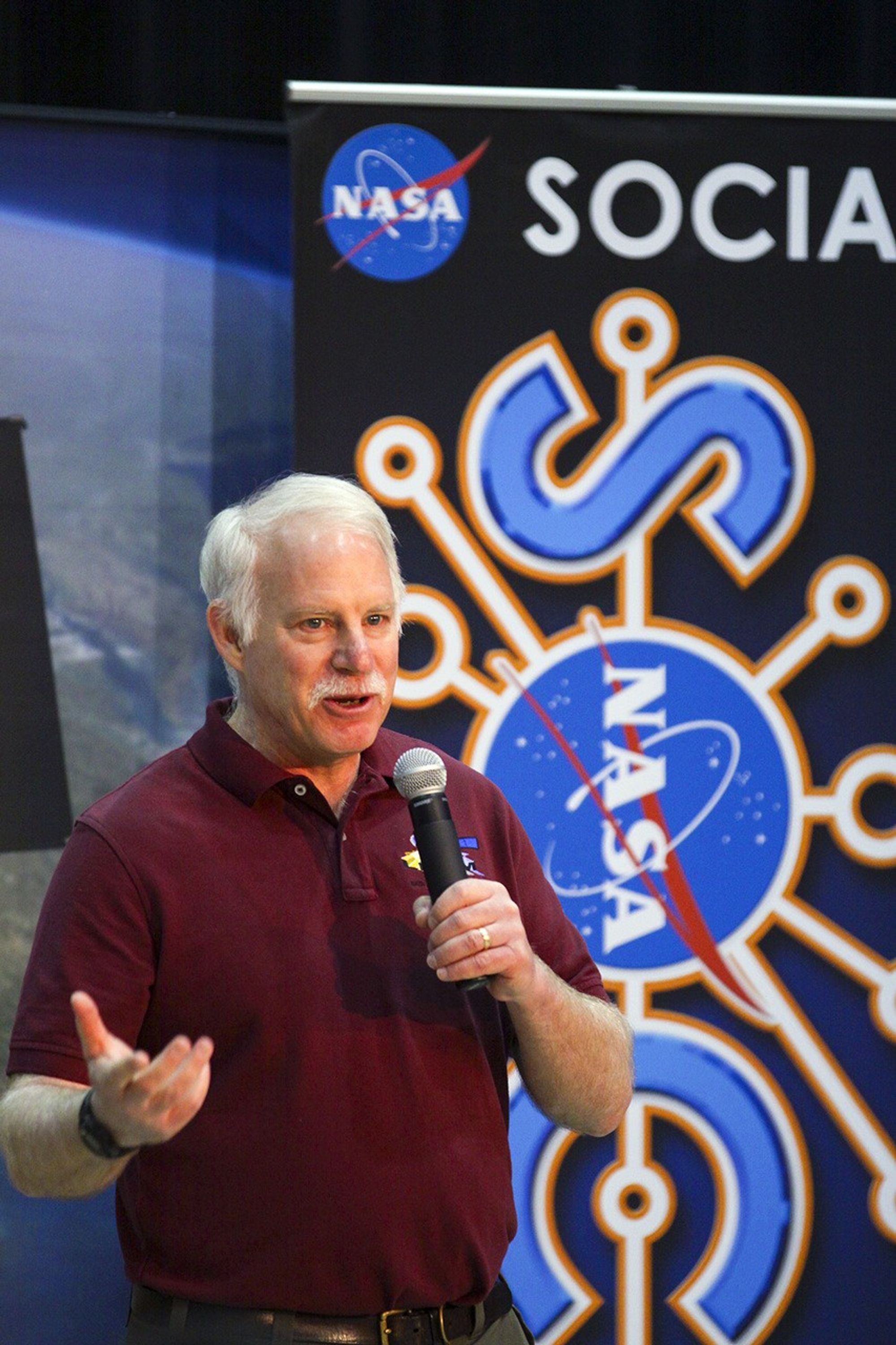 A man is slightly off center in the image. He is looking to the right of the camera and is holding a microphone in his left hand, looking to be standing and presenting or making a speech. He is wearing a maroon, short-sleeved polo and has white hair and a white mustache. Behind him, in the background of the image is a banner with the NASA meatball logo and a logo for a NASA Social.
