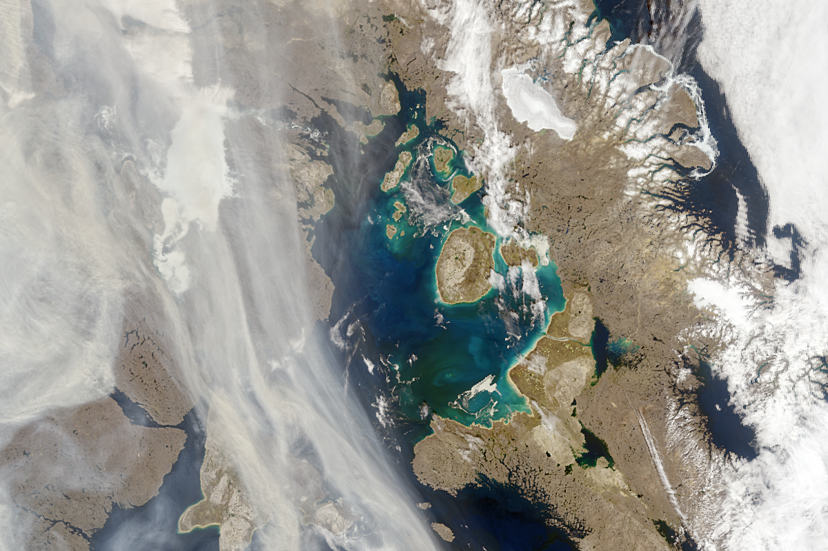 A satellite view of land, water and smoke is seen. Land covers the top, left, and right portions of the image, and is shown as a light brown. The right side of the image is splotched with white snow. Interspersed between the land is ocean water, usually in a deep blue color, but sometimes swirling with blues and greens near the shore, showing sediment. On the left side of the image, wisps of light brown smoke glide over the land in a motion from top to bottom.