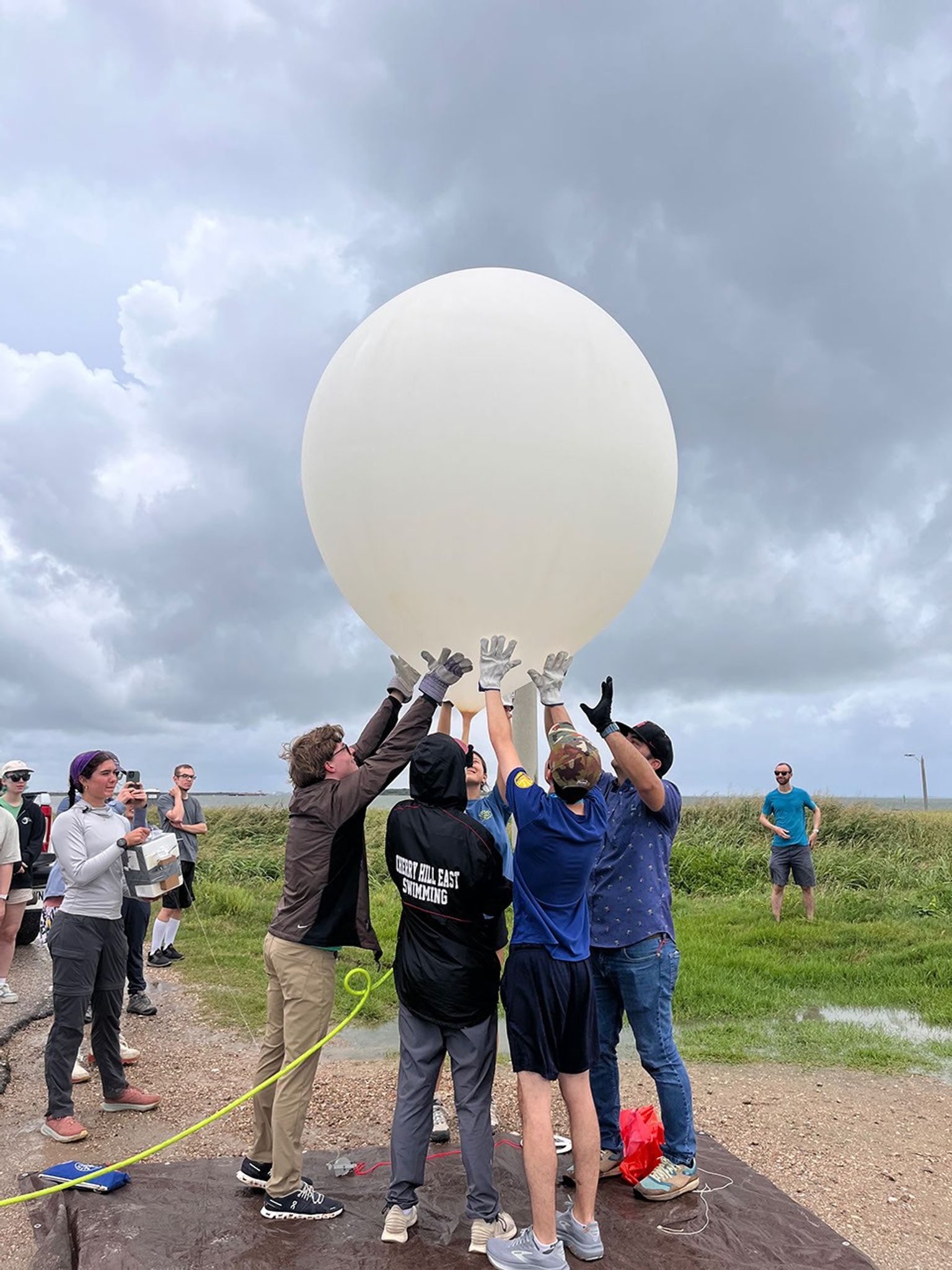 The center of the image shows a large white balloon. Beneath the balloon are five people in a circle, gently holding at the bottom of the balloon to keep it upright. In the background of the image, other people watch.