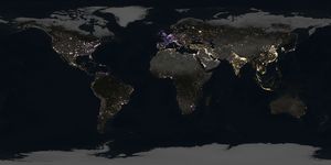 Gold, purple, and white points of light brighten up a globe shrouded in darkness, while a shaft of sunlight is just visible peeking over the edge of the planet.