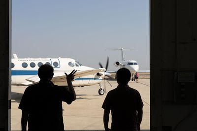 Two men stand silhouetted inside an airplane hangar, while a twin-propeller NASA aircraft takes off in the background.