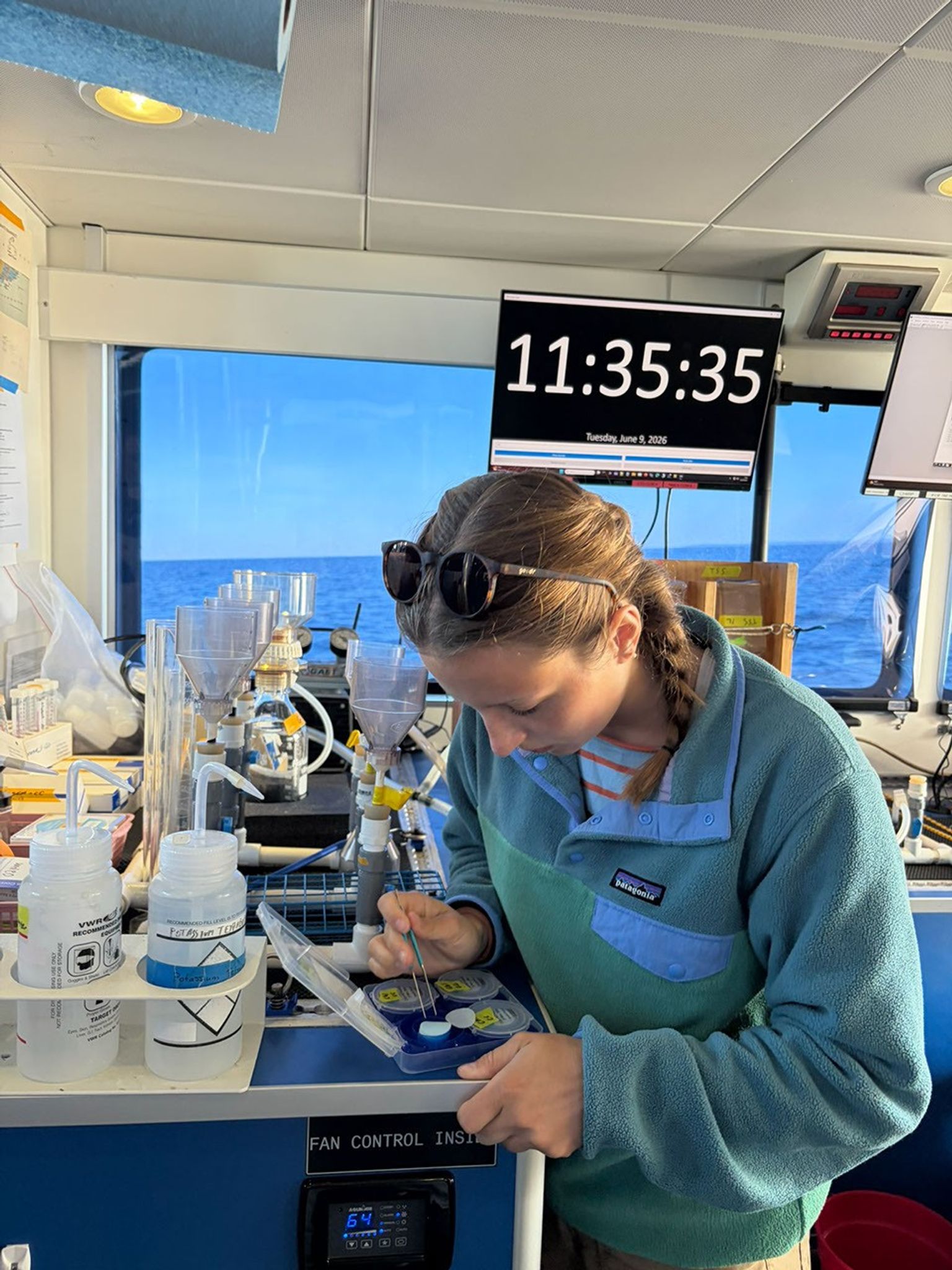 A young woman wearing a fleece top, with sunglasses on her head, bends over a lab bench, using a tweezers to adjust a disk of filter paper. In the background, a window gives onto the sea.