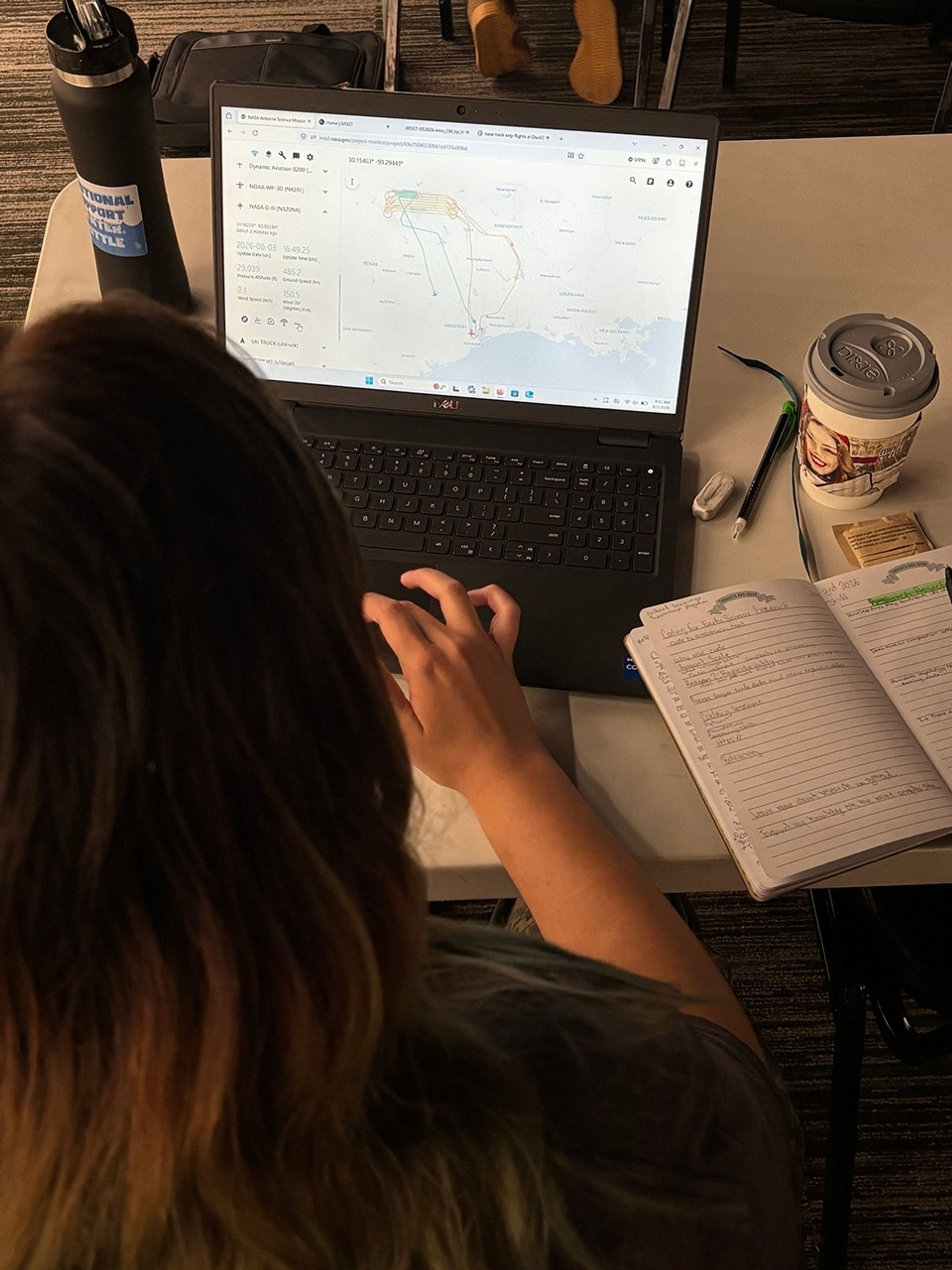 The image is taken from above a student working on her computer. On the bottom left of the image, the back of the student’s head is shown. Beyond her, a laptop is open to a map with lines highlighting the flight routes.