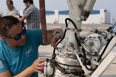 A woman kneels beside the landing gear of a large airplane, looking at it closely.