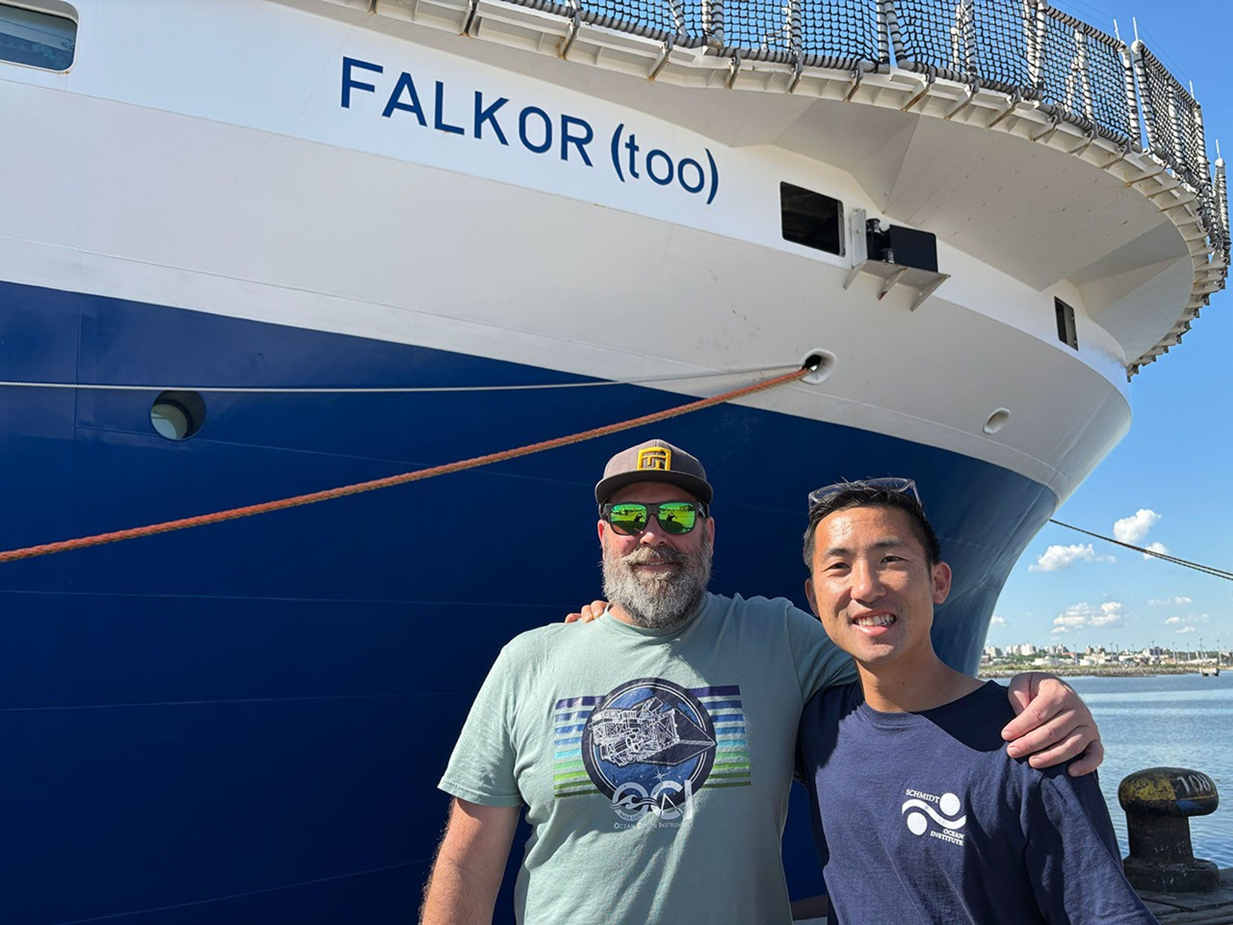 Two scientists pose on the dock beside the research ship, with its white superstructure and deep blue hull. In the background is a shoreline: a low city of white buildings fronted by a rocky quay.