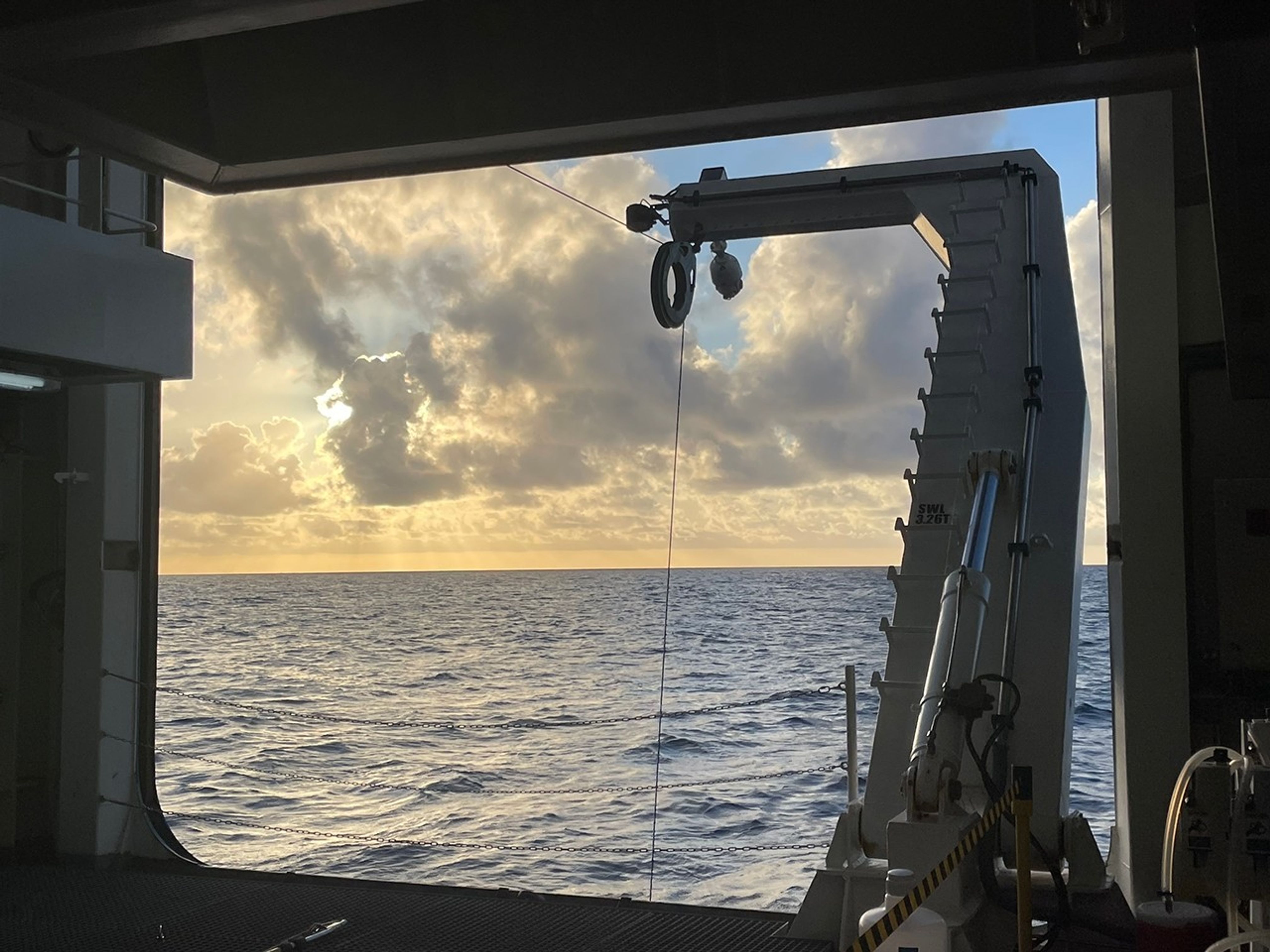 Under a sunset sky, we view the ocean through an open bay. The view is bisected by a cable extending from a pulley atop a crane into the sea: the CTD (conductivity, temperature, and density) water sampling rosette is at work below the surface.
