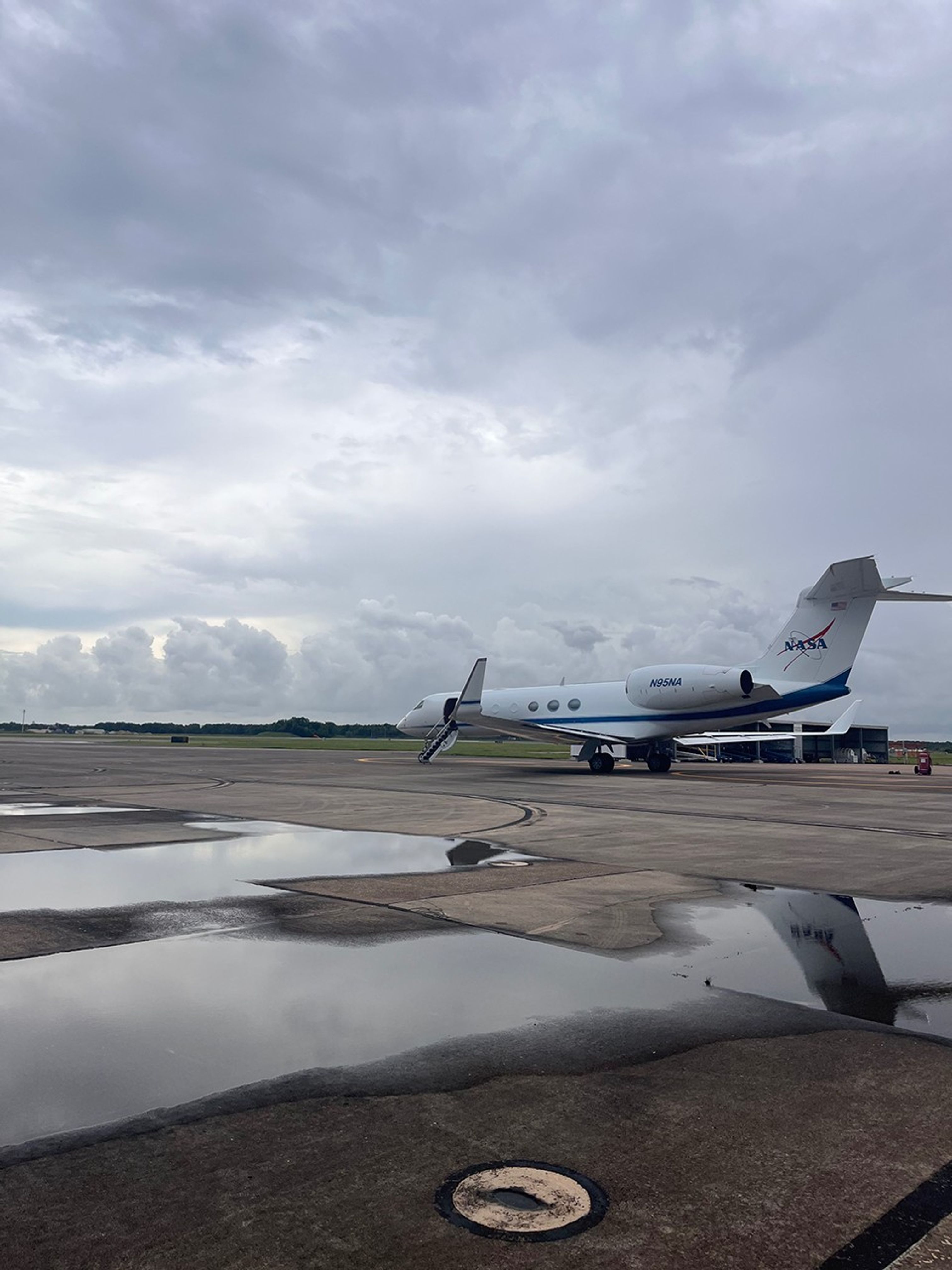 A white aircraft is centered in the image. The background is sky is full of wispy white and dark gray clouds. The foreground ground is a concrete runway with puddles of water reflecting the sky and tail of the aircraft.