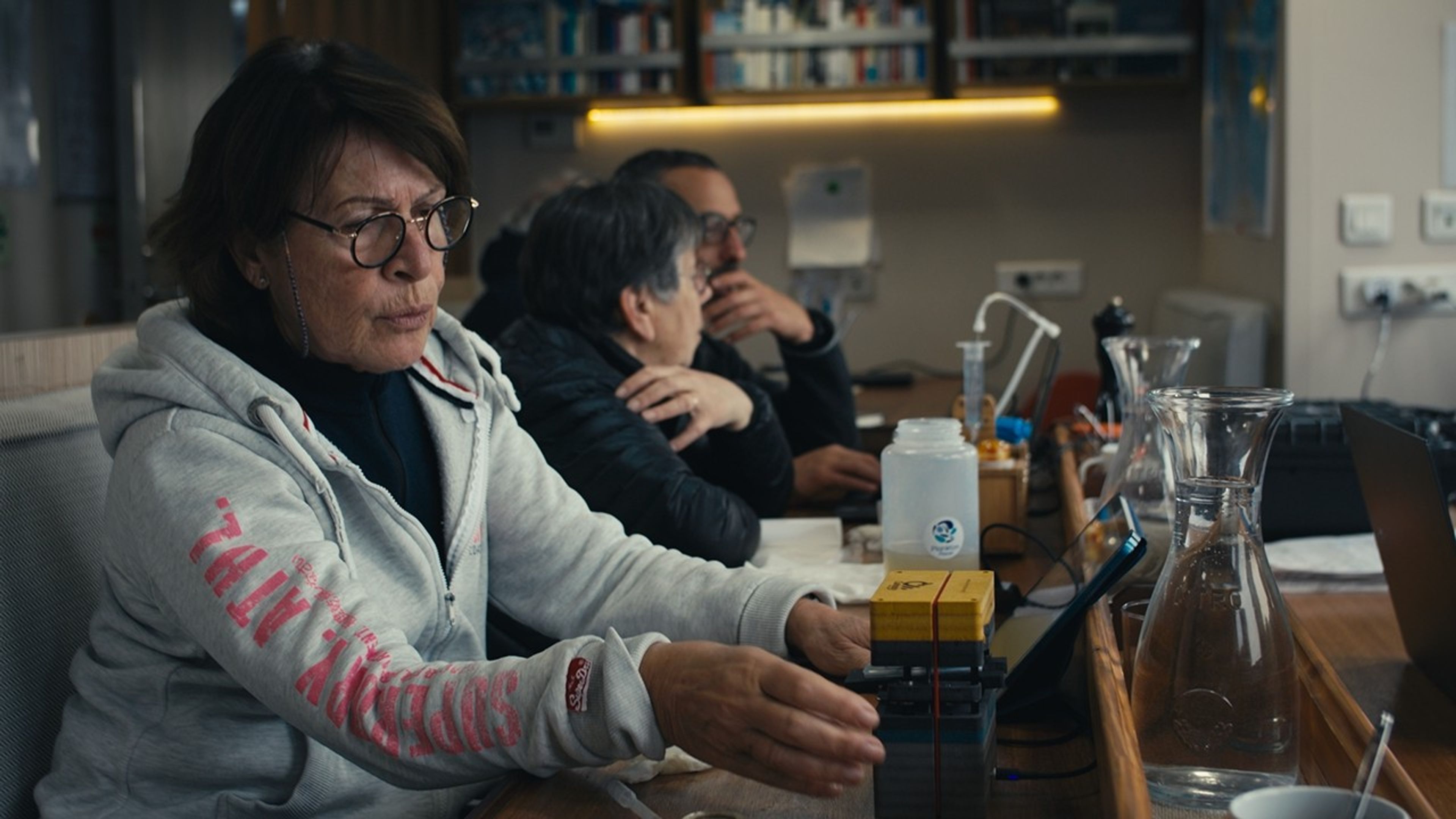 This interior shot shows three lab workers in a science lab. On the bench are several flasks of water, a yellow and black box, other tools that are partly blurred. In the foreground, a woman in a turtleneck and hoodie seems intent on something in her hands that we can’t see; to the rear, a man and woman are in conversation, their eyes on something on the bench.