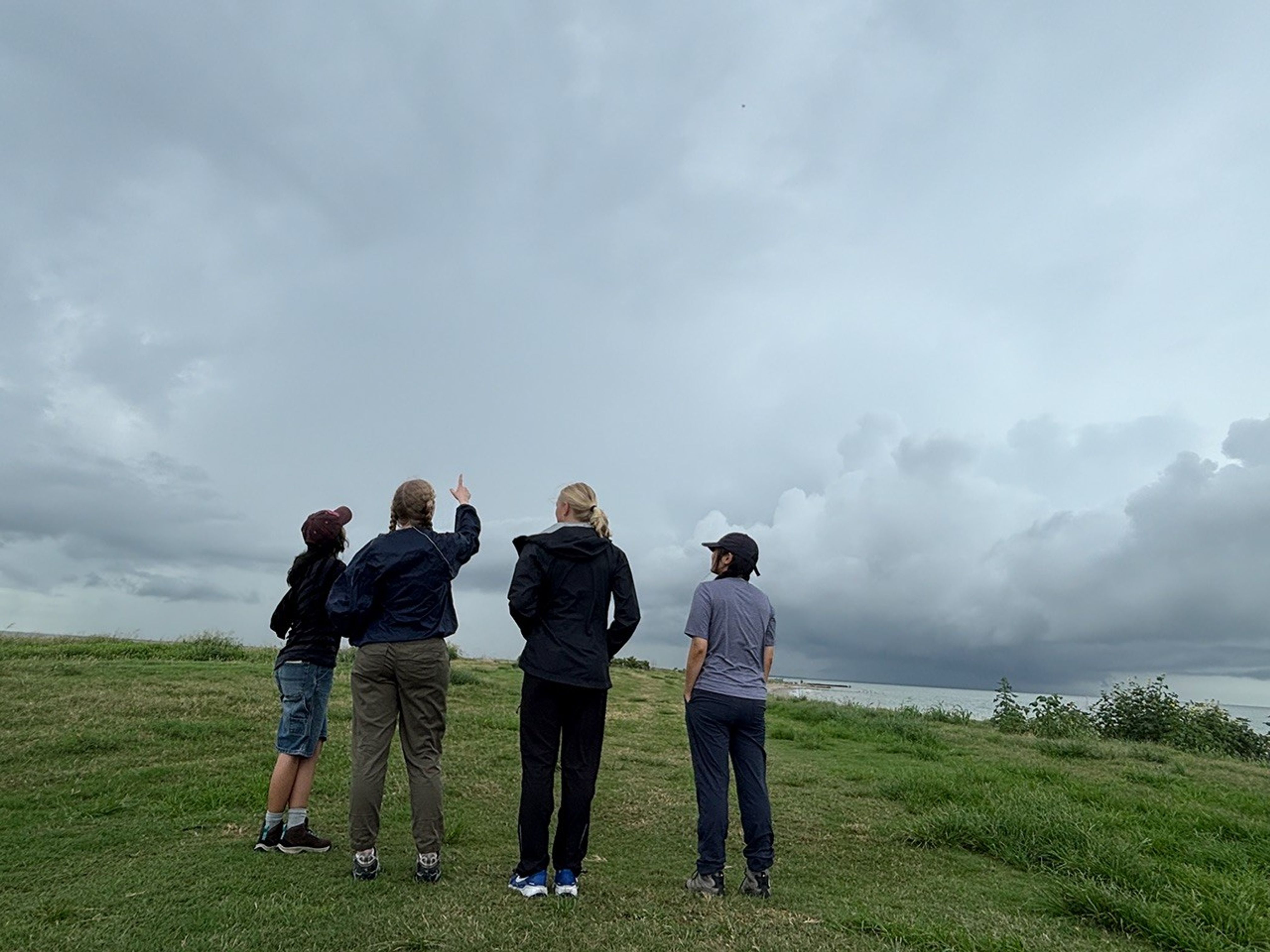 Four students stand in a line centered in the image facing away from the camera. They’re looking up at the sky — a dark and cloud filled sky — and standing on dark green grass. One student, second from the left, points up at the sky.