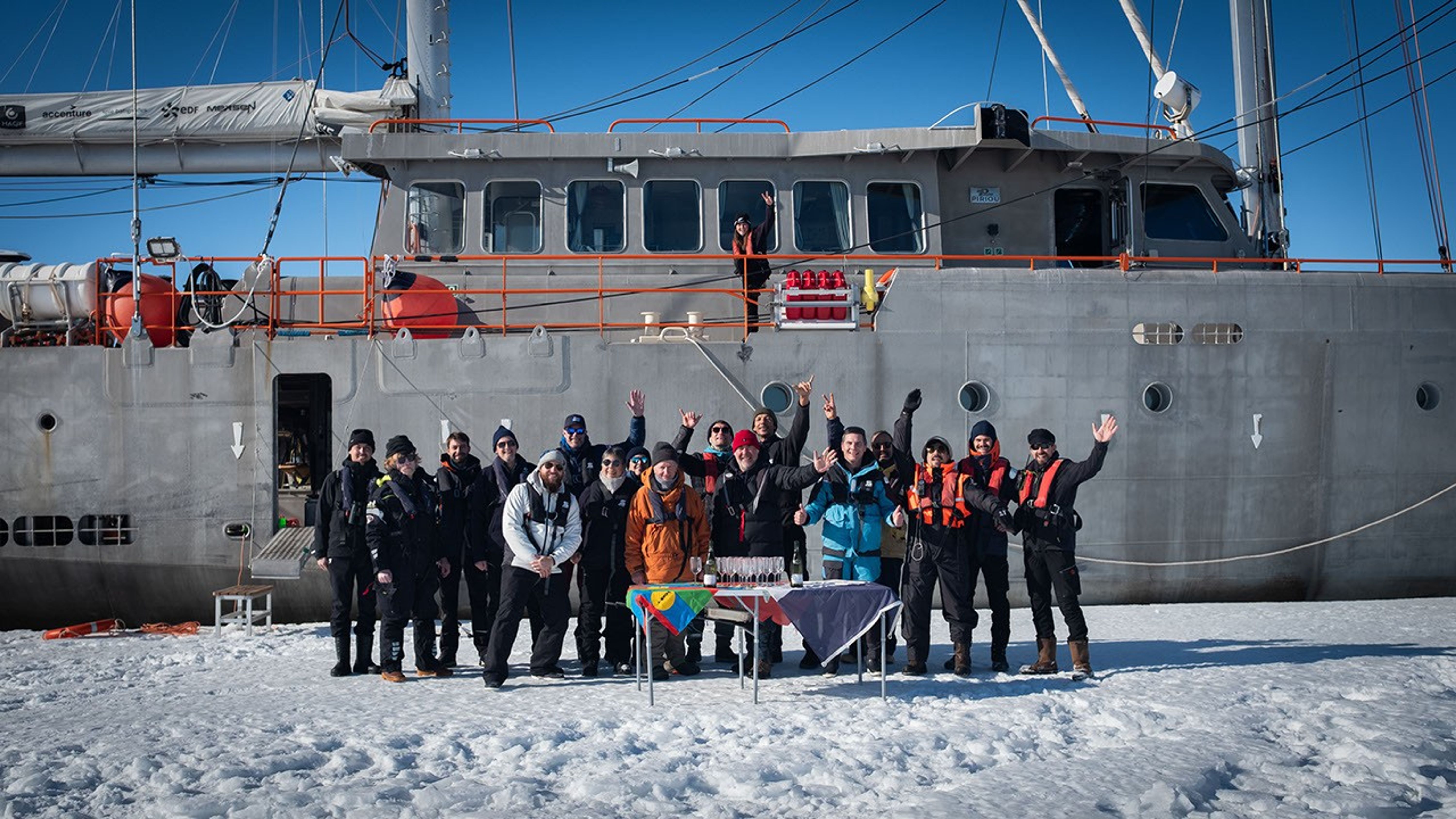 The gray hull of Perseverance, her sails furled, forms the background of this photograph. To the left, a door in the hull is open, and a gangplank extends down onto an ice floe. There we see the entire crew (except for one sailor visible in the window of the ship’s bridge) posing and waving behind a folding table set with bright tablecloths – and the French flag! – and wine glasses and bottles. Everyone is dressed for the cold, and several are wearing life vests.