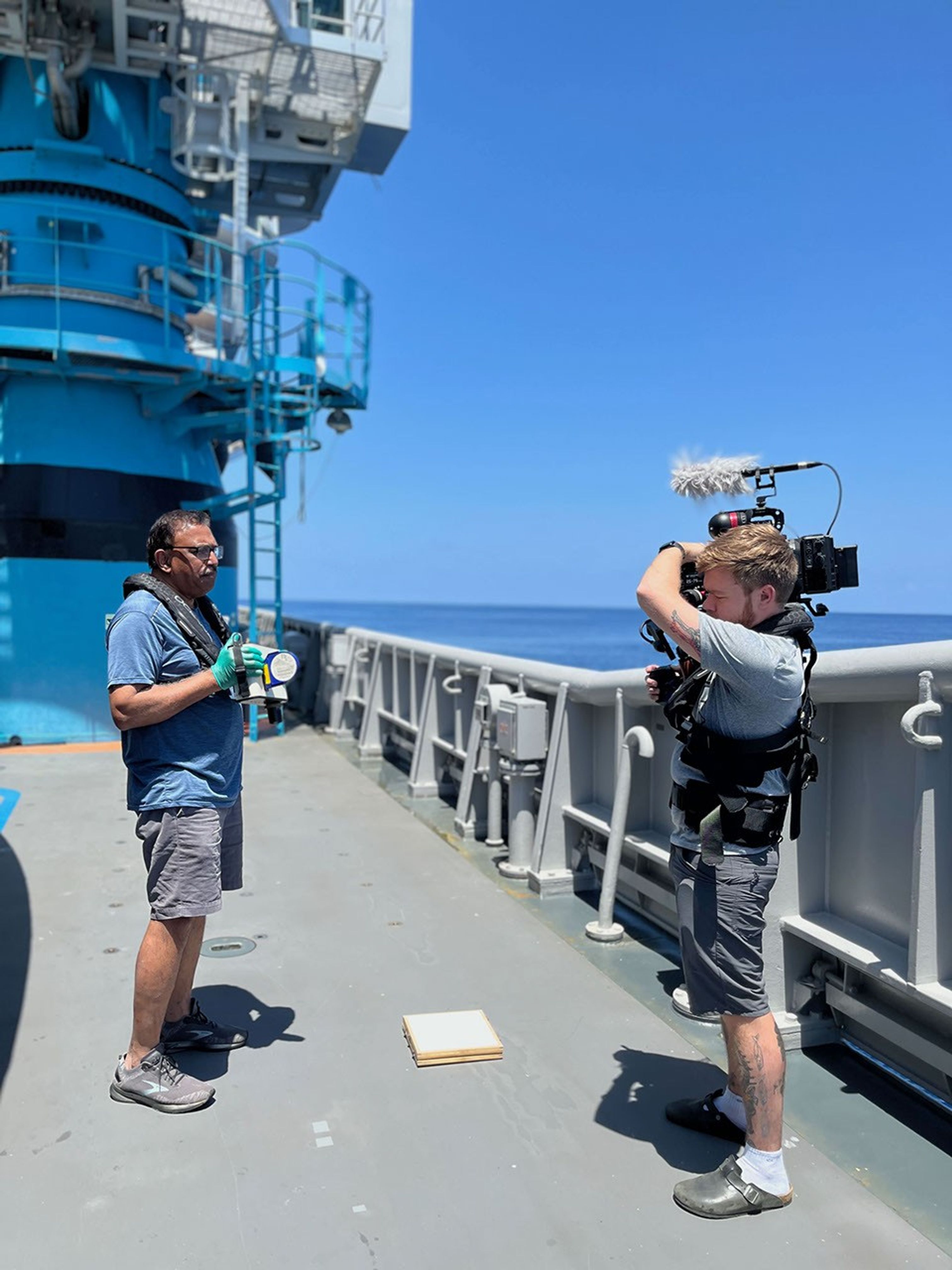 Two men stand on the deck of the ship. To the left is a middle-aged man with a safety vest over a t-shirt, shorts, and sneakers. He holds a pale gray and black in his teal-gloved hands, showing it to the other man, also in a safety vest, with a film camera on his shoulder. It’s a bright, sunny day with a calm blue sea in the background.