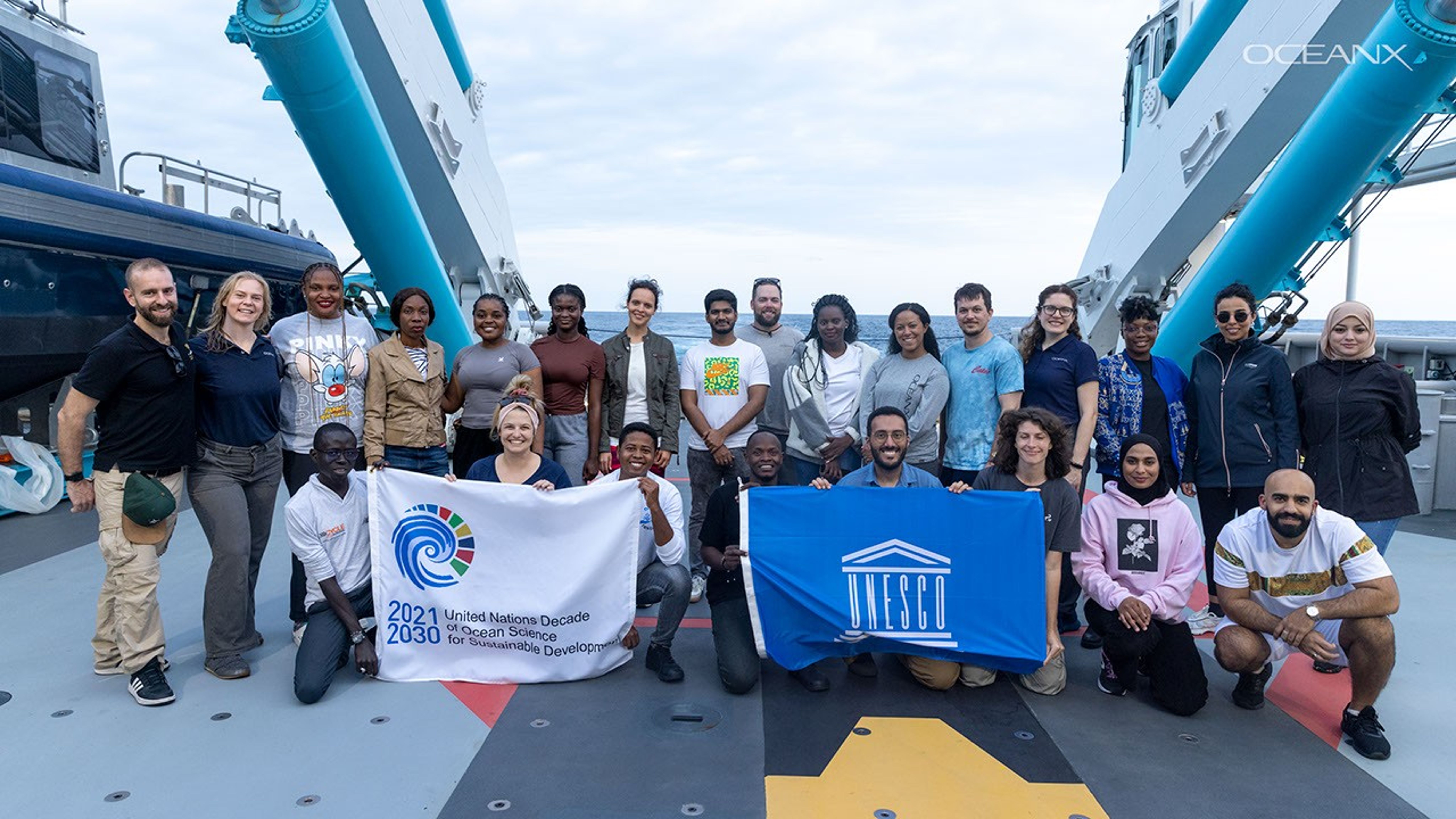 In front of the bow of the elegant white superstructure of the ship, the OceanX group poses for a formal team shot: a multiracial group of young to middle-aged humans. They display the dark blue flag of their expedition, with the word OCEANX in white.  