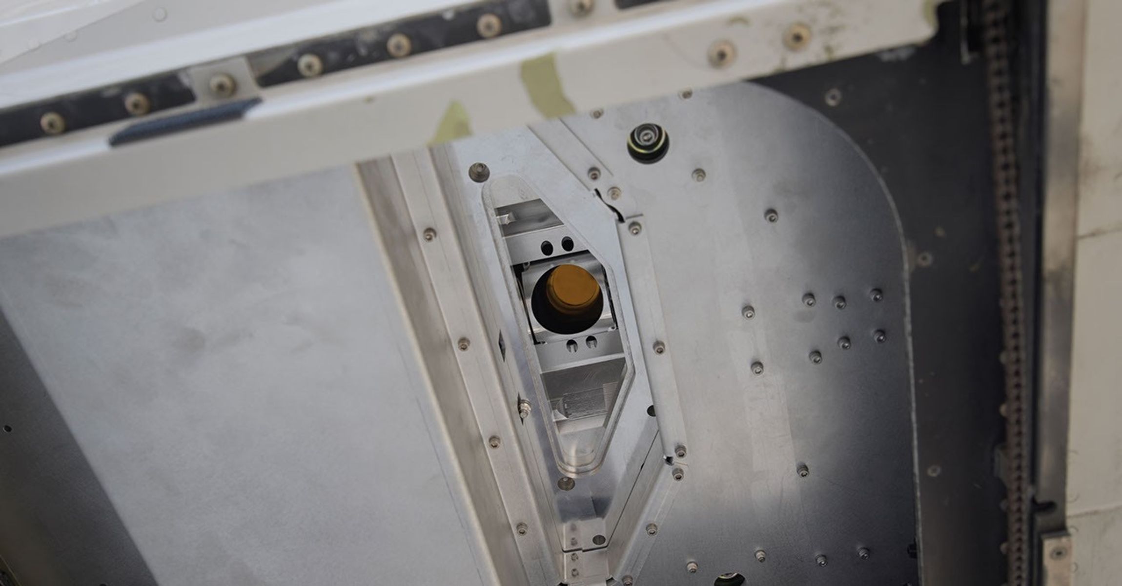 A copper-colored lens looks down from a narrow port beneath an airplane.