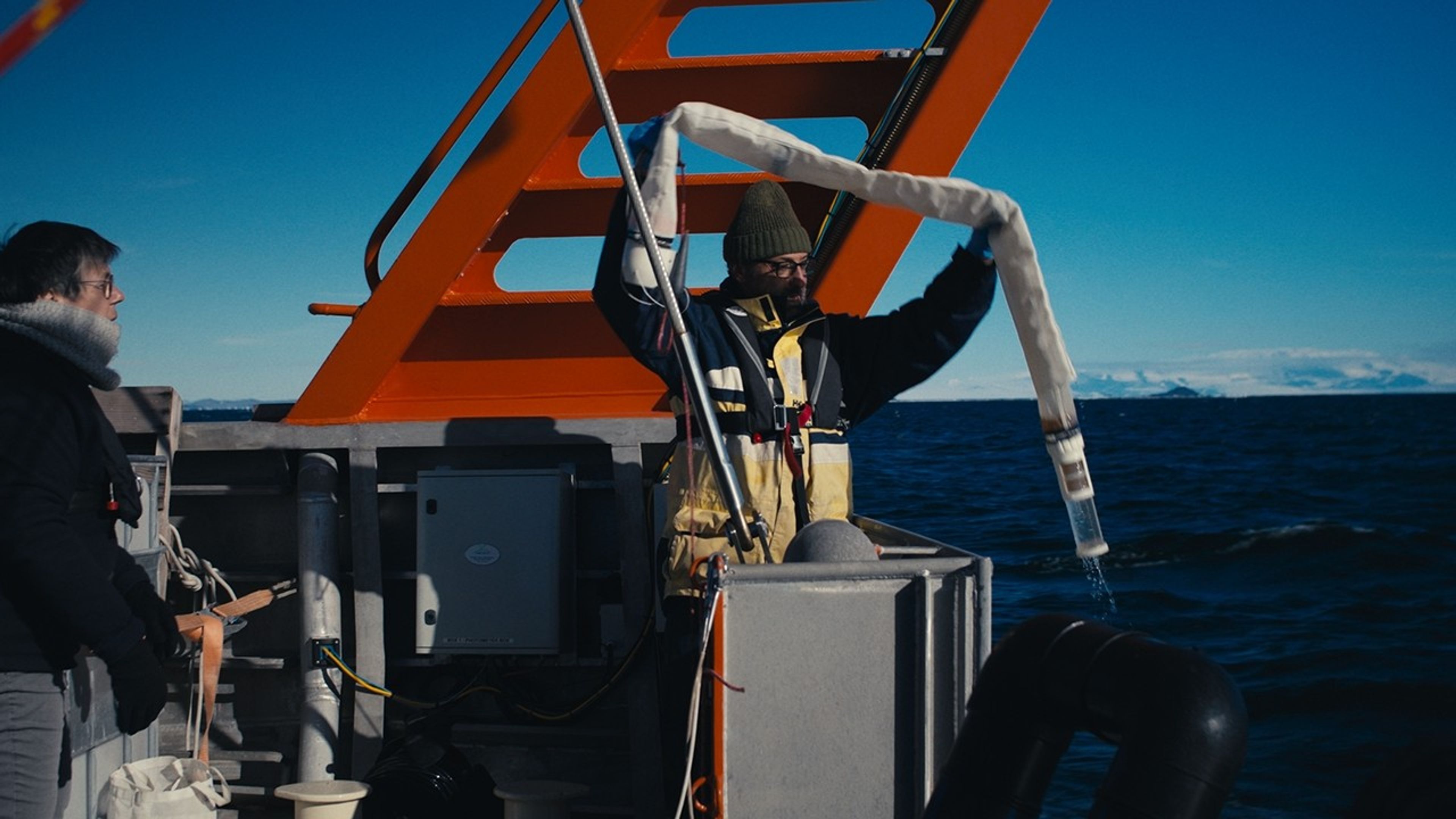 The bright red superstructure of the ship frames the scientist, dressed in a parka and knit hat, who stands holding the long “snake” of a plankton tow just retrieved, over his head. To the right, the bottom of the net extends into a collection bottle. He’s ready to examine what’s inside. Behind him is the gray gunwales of the ship, a deep blue sea and, on the horizon, distant snow-covered mountains. Another scientist stands by partly out of frame, waiting to assist, collar turned up, bucket in hand.