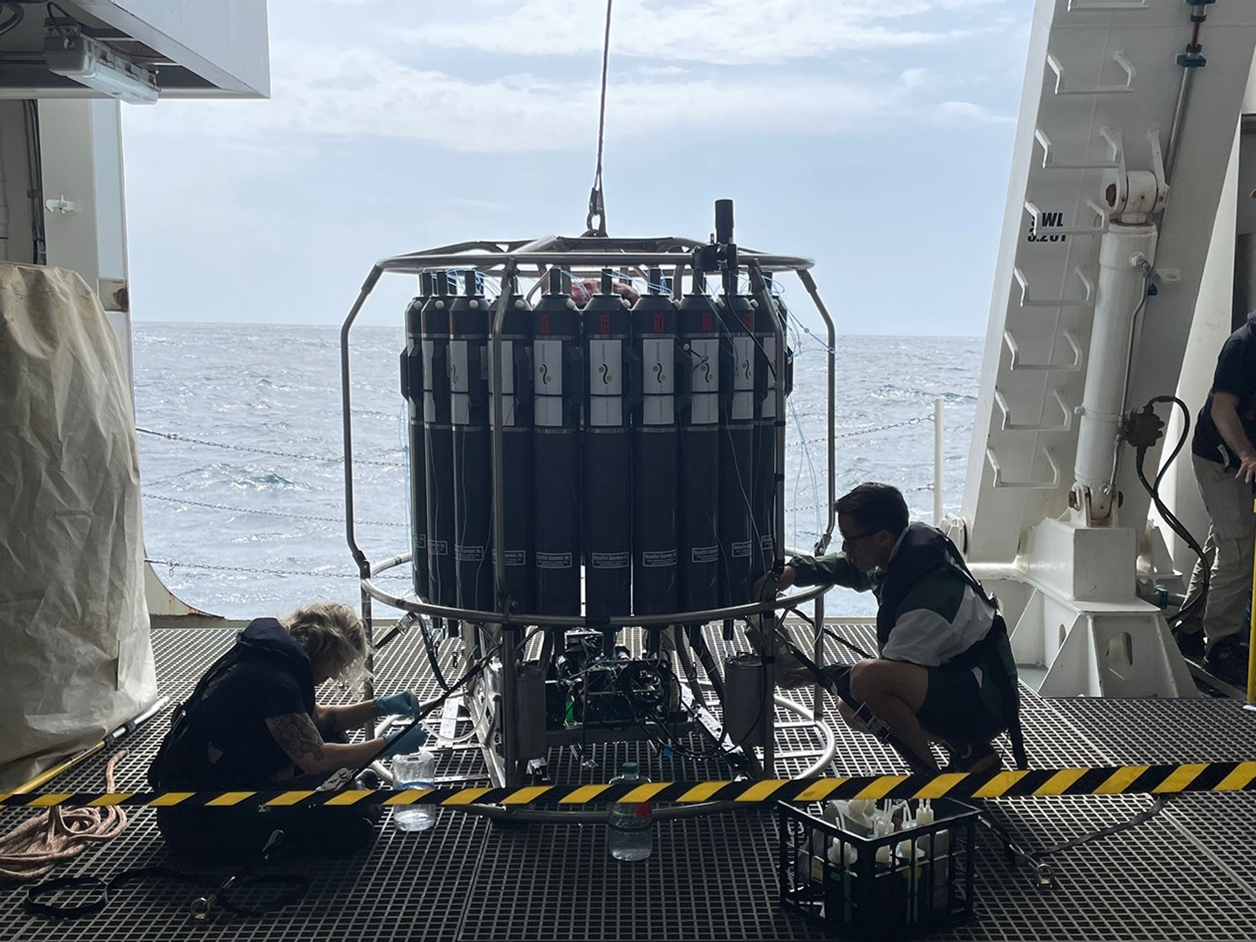 Two scientists squat on the grated ship deck, behind a caution tape. They are collecting samples from the 24 tall gray Niskin water bottles that fill the circular steel frame of the water sampling rosette. In the bottom section of this device, below the water bottles, we can see instruments used to collect data through the water column. Thin cables are triggered at the appropriate depths to close the valves of the bottles. At the top of the rosette, a heavy clip connects to a cable suspended from a crane that is unseen above; this will be used to hoist the rosette in and out of the ocean.