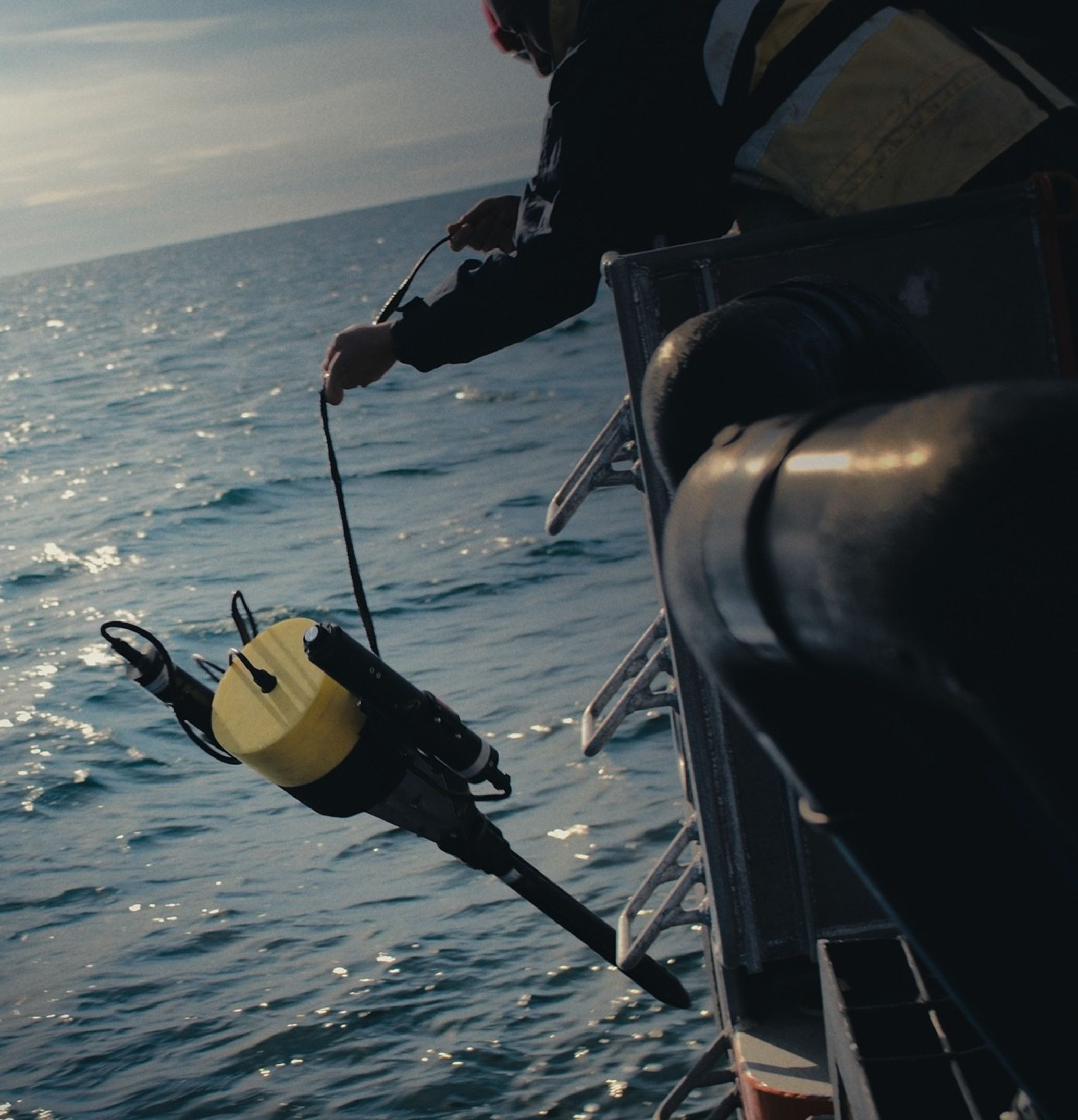 A person holding a line with both hands suspends a yellow capped device with two cylinders on opposing sides, all connected by wires, over the side of a boat. A pointed shaft extends from the bottom of the device, open water is on the left side of the image and the body of the boat on the right. The person is largely cropped from the image at the top right, they are wearing long sleeves.