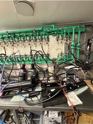 A lab bench contains and assortment of black cables, hoses, and pumps.  Behind it, against the wall hangs a system of bright mint-green pipes and valves with orange and red circular covers.