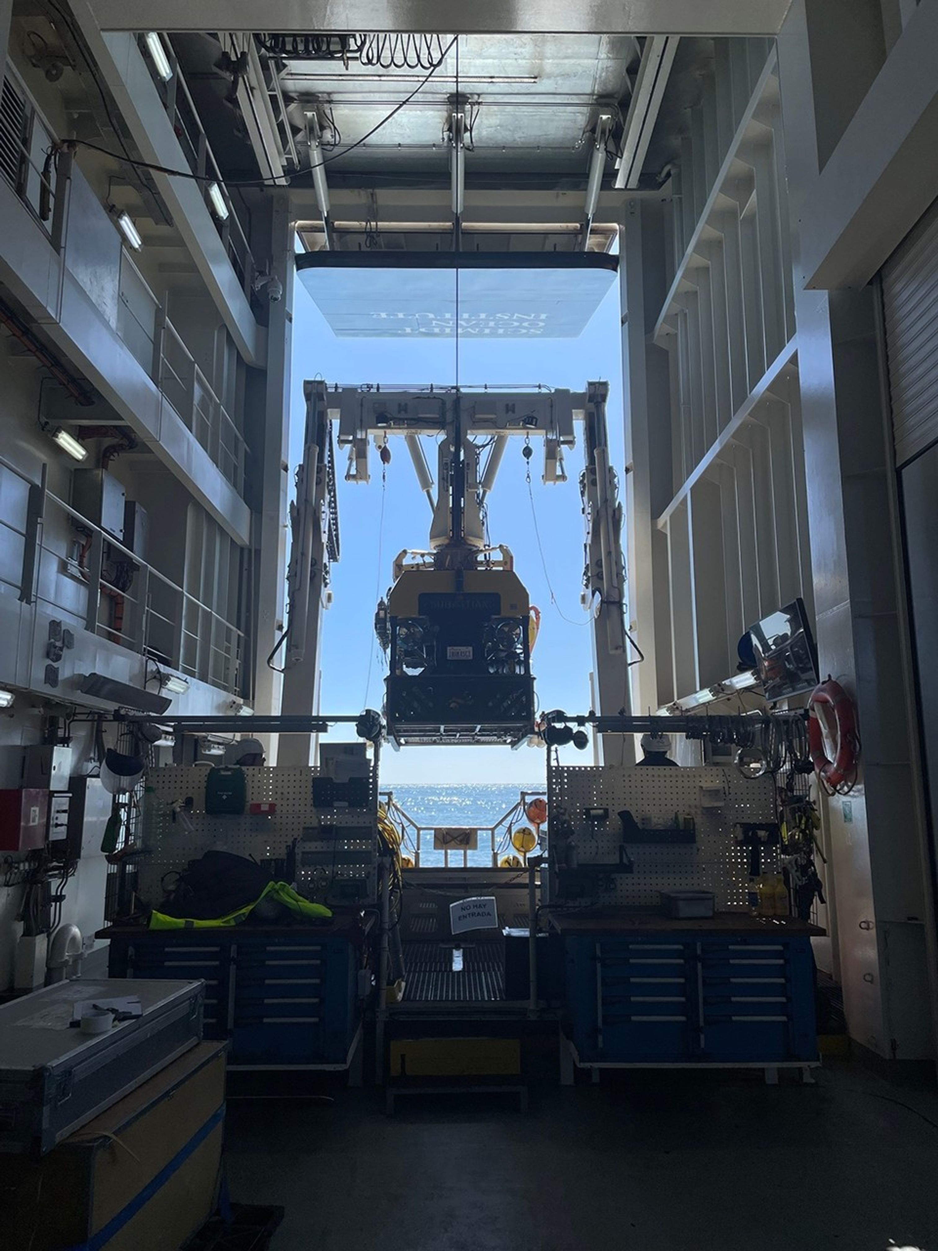 We look through the dim interior of a shipboard hangar to a remotely-operated vehicle, suspended from the ship’s A-frame and waiting to be lowered into a sparkling sea.
