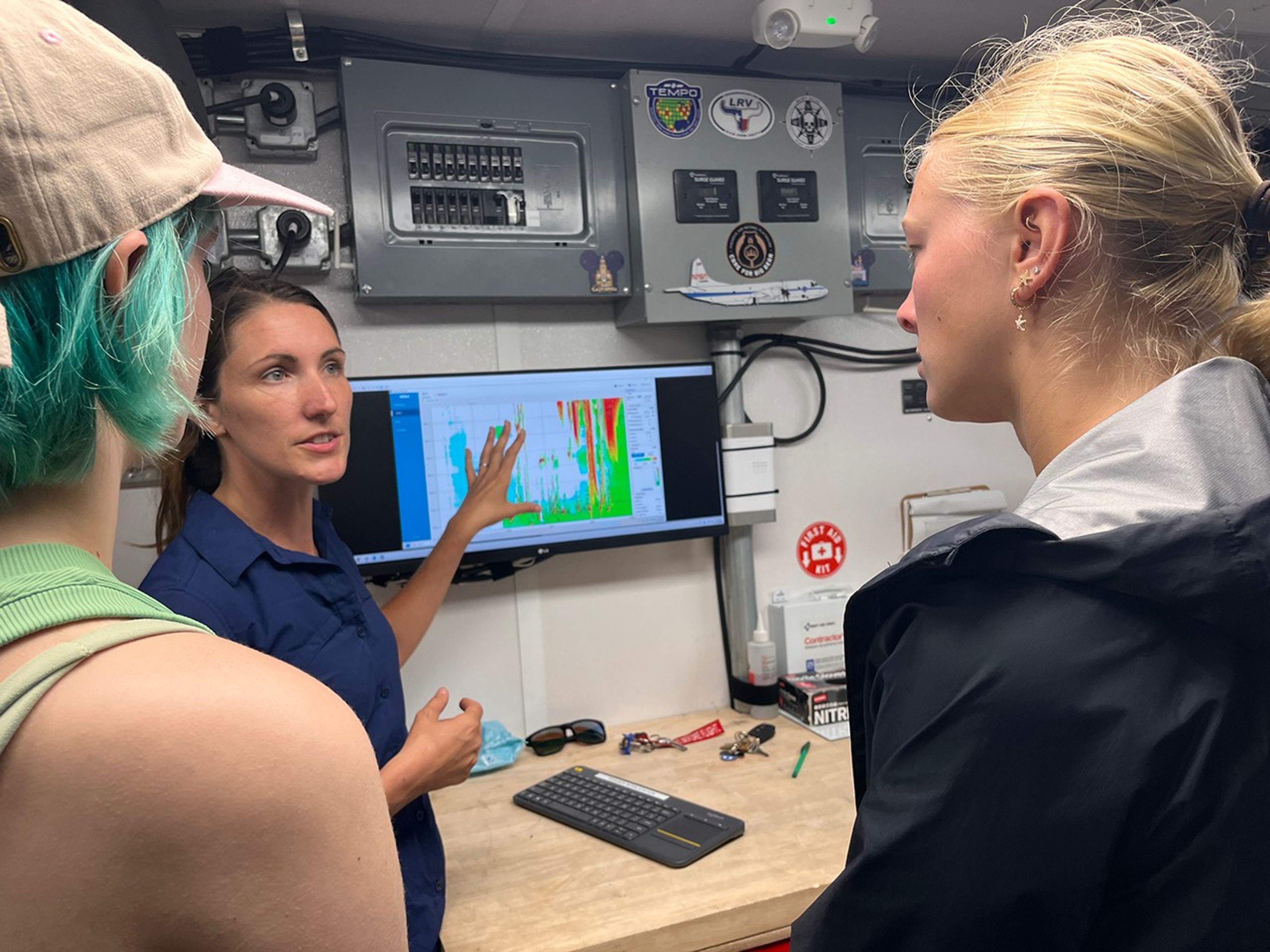 In the background of the image is a computer screen showing science data in green, orange, and red colors. Two students, in the foreground of the image, are looking at the screen, while a woman stands in front of it, looking at the students and pointing at the screen.