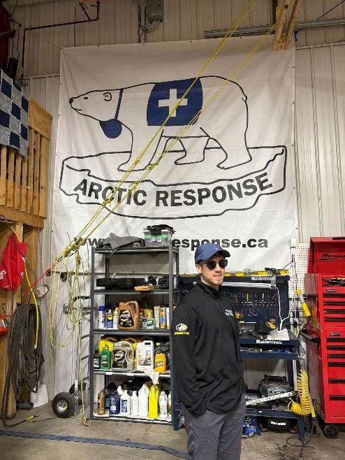 A young man in outdoor clothes stands among shelves of supplies. Above is a giant poster of a polar bear wearing the symbol of the blue cross, with the logo ARCTIC RESPONSE. Arctic Response (or Arctic Response Canada) is a safety and rescue organization.
