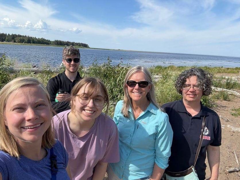 The team of researchers takes a selfie with the lake in the background. They’re wearing short sleeves and sunglasses, and smiling broadly.