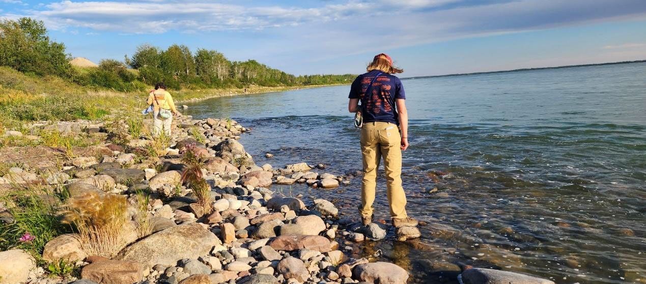 Two researchers hike along the pebbled edge of a shallow river. The river stretches into the distance to the right; to the left, stands of deciduous trees in full leaf provide the background to a riverside meadow of tall grasses and other plants.