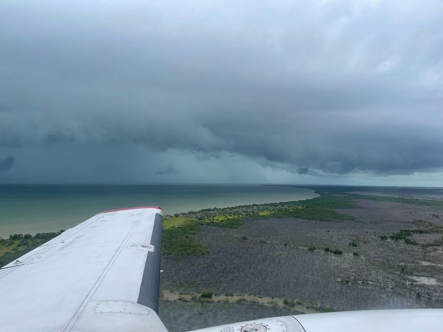 The plane’s wing is shown flying over land that is primarily a dead gray, with occasional spots of green vegetation. The sea on the horizon reflects a sky of stormy clouds, with the water shading from pale green at the coast to deep blue-gray where sea meets sky.