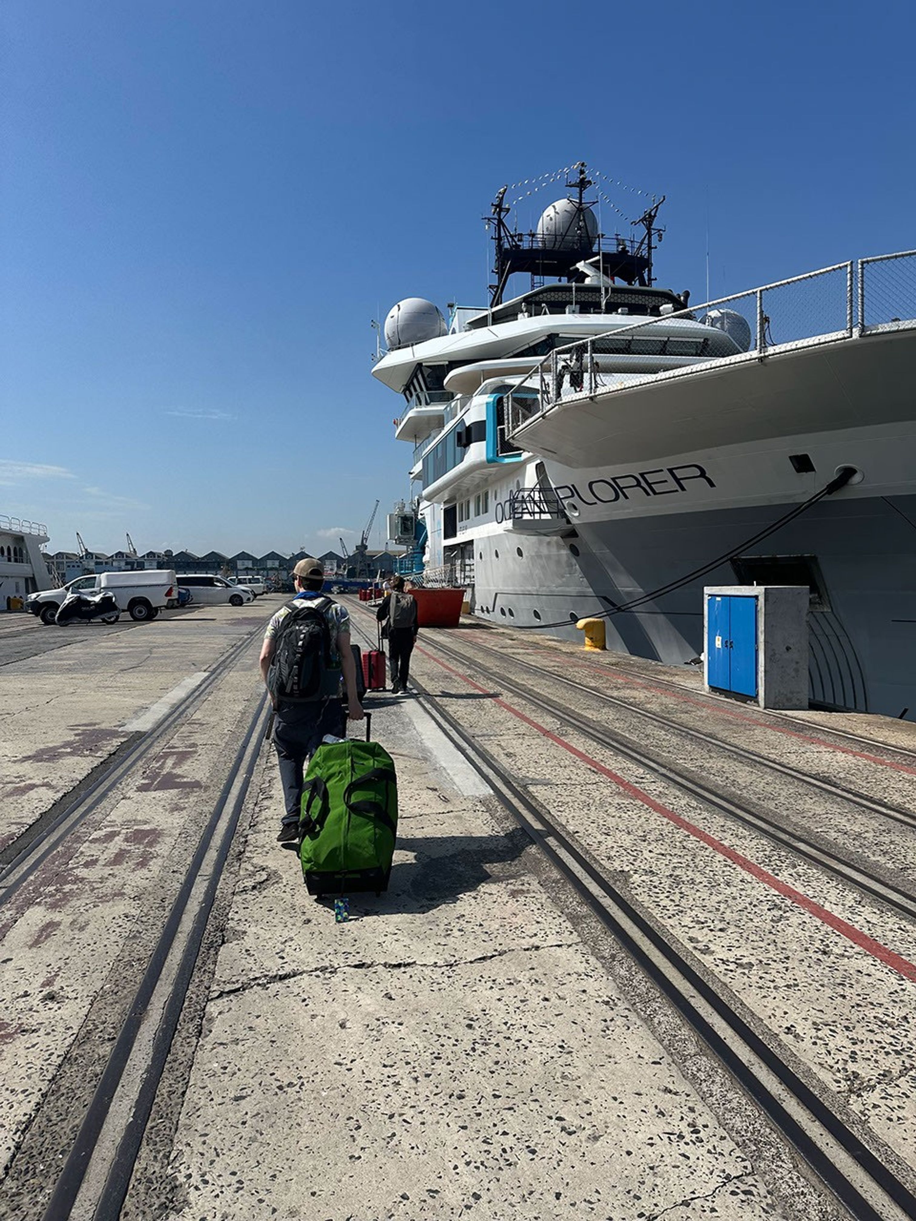 We see two expedition joiners from the rear as they tow their luggage along a concrete wharf lined with tracks for cargo. To the right, the Ocean Explorer ship rises above their heads – a gray-hulled research vessel with a white superstructure, topped by antennas. In the distance stand low buildings and parked vehicles.  