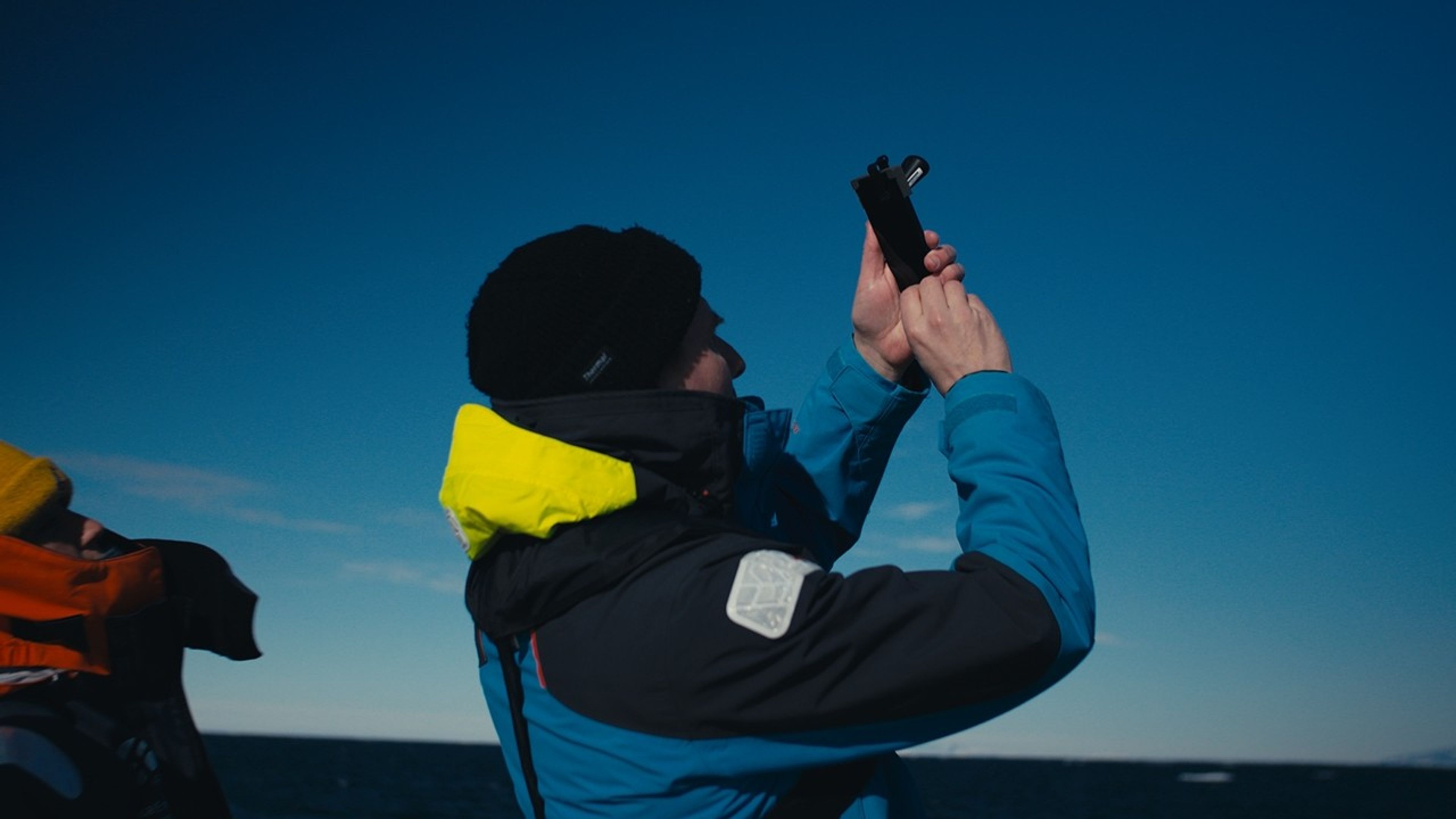 This is a profile view of a man in all-weather gear and a cold-weather hat, holding up a photo toward the sky. 