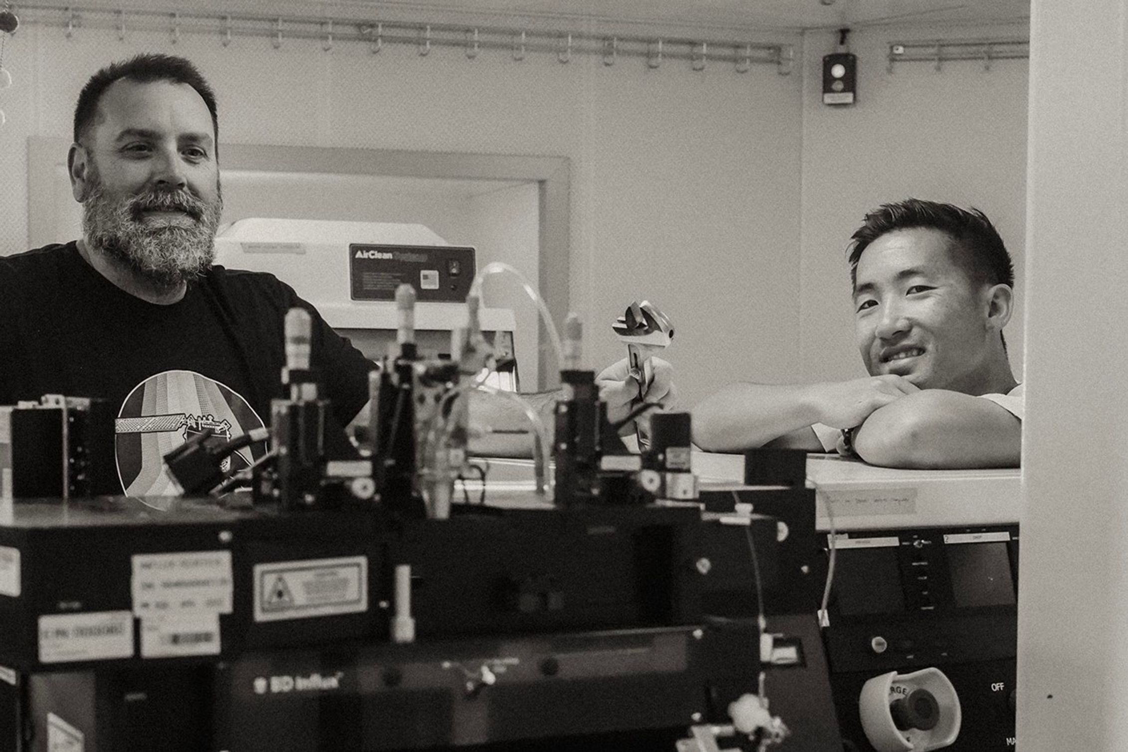 A black, blocky device accessed through tubes and coils rises to chest height on the two smiling men shown. The background is a shipboard laboratory with a large screen display.