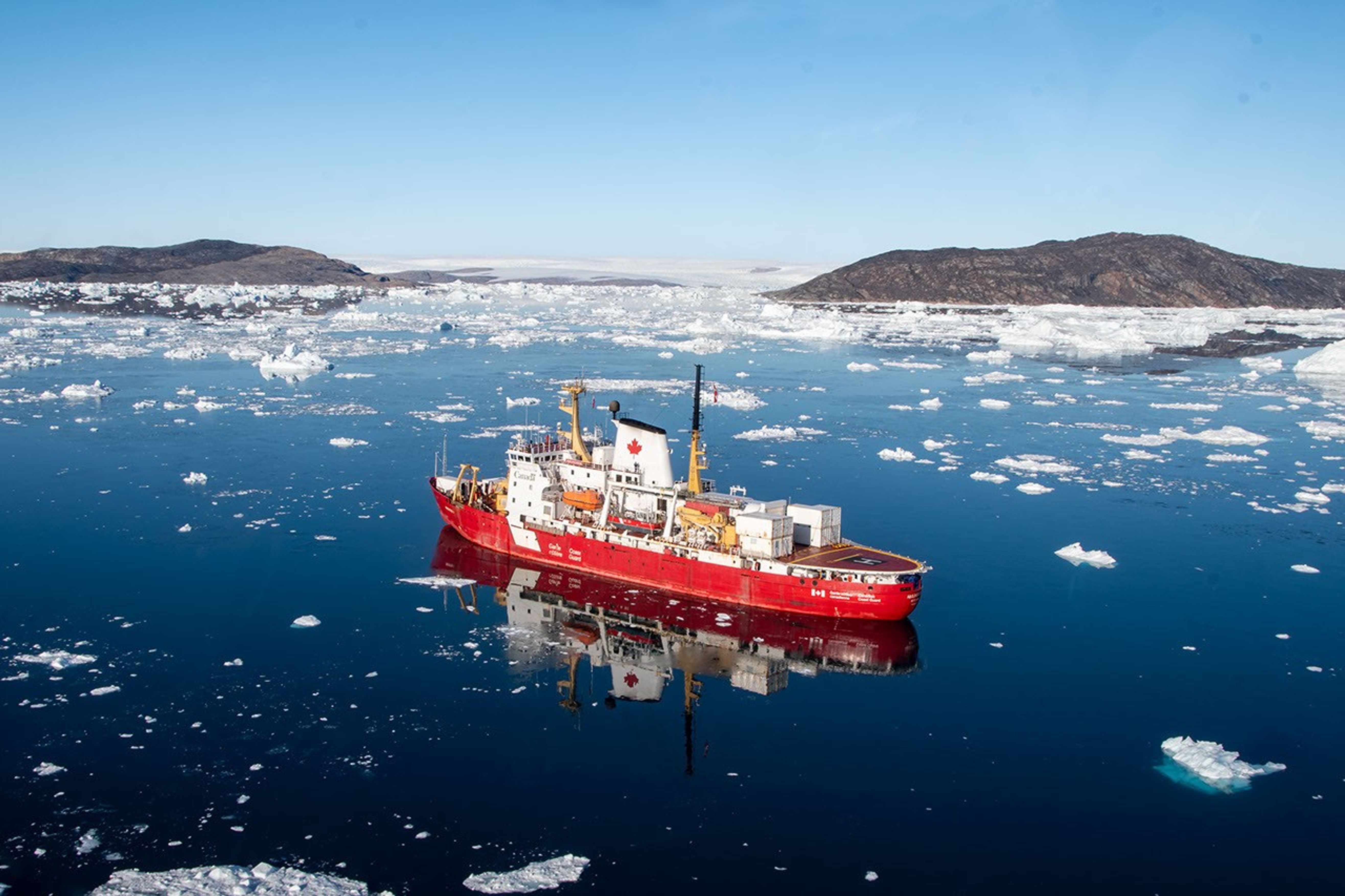 A red-hulled research ship with a white superstructure boasting the Canadian maple leaf sits on a glassy, bergy-bit-strewn sea with bare hills on the horizon.