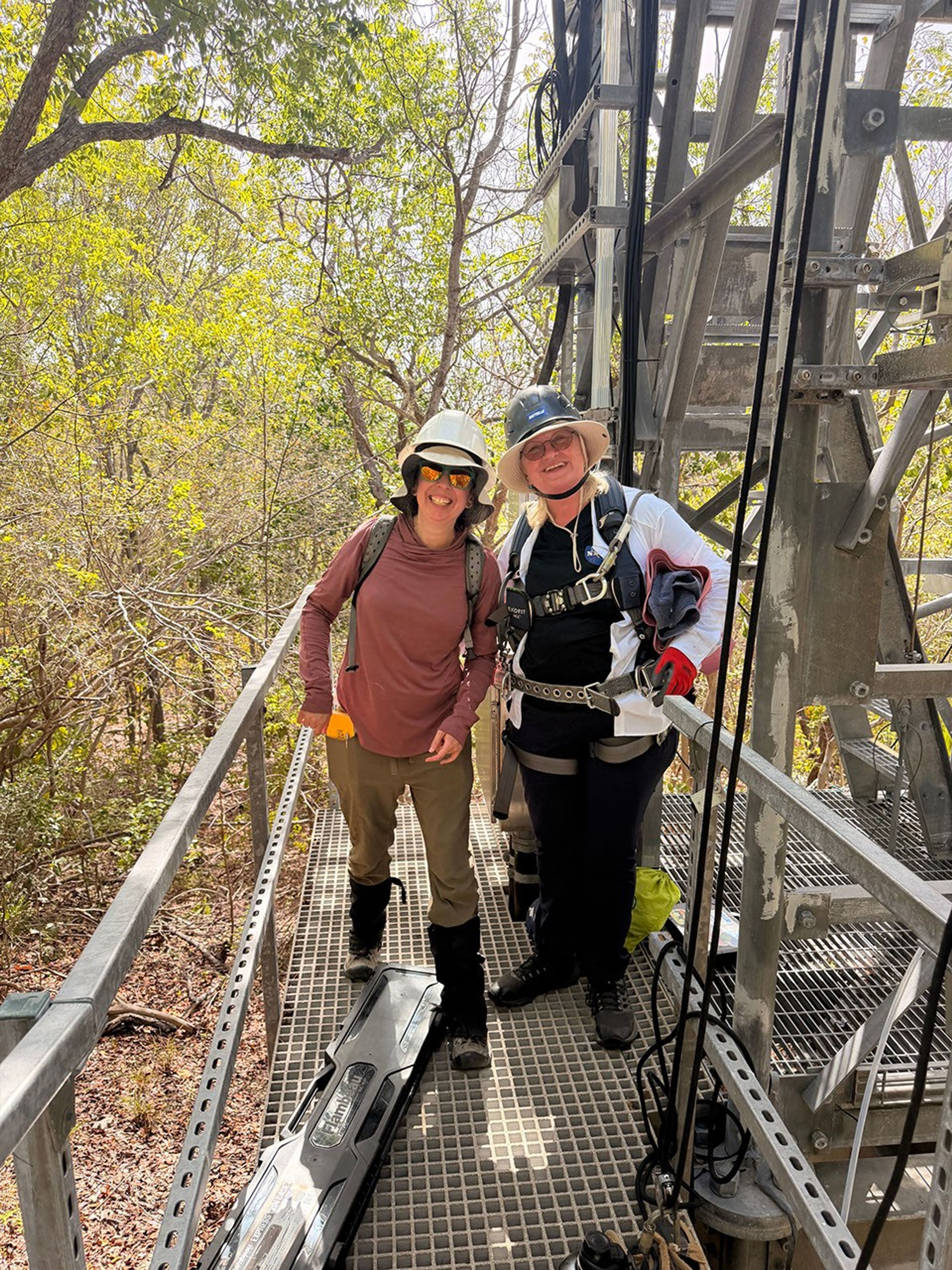 Two women in outdoor gear, heavy boots, and hard hats stand on the grated low level floor of an observation tower.  