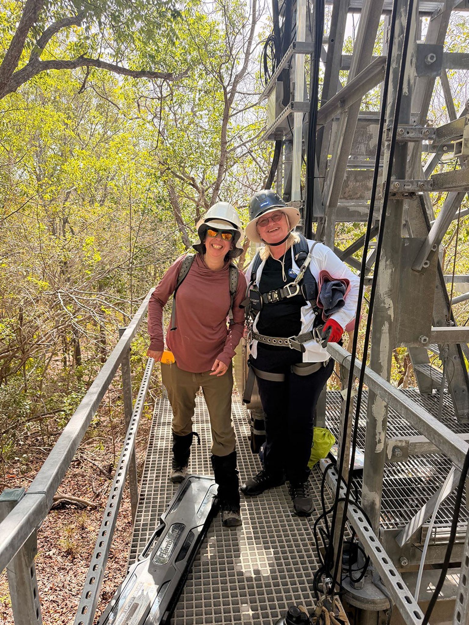 Two women in outdoor gear, heavy boots, and hard hats stand on the grated low level floor of an observation tower.