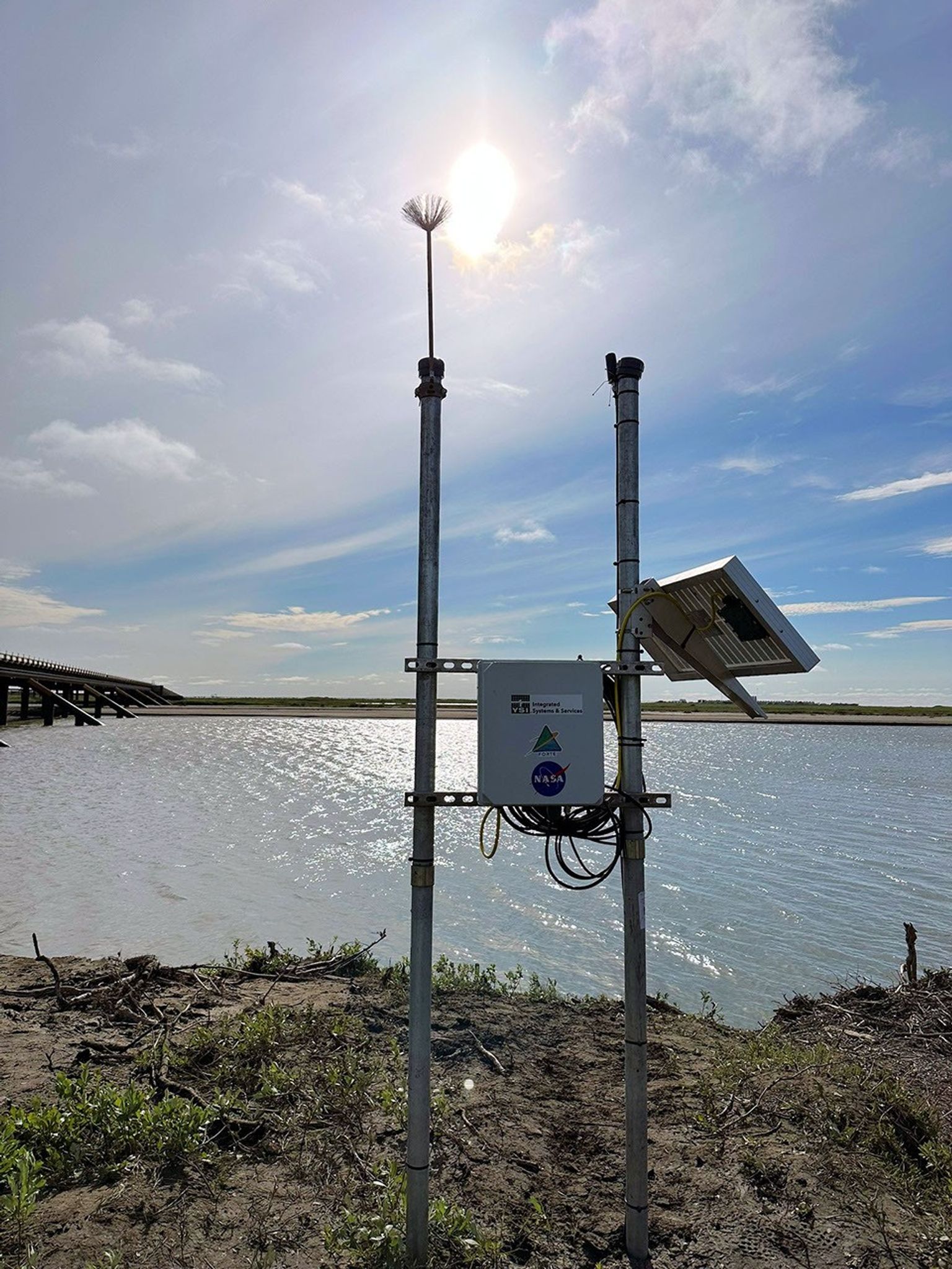 Two poles driven into the sandy dirt at the edge of a river are connected by two crosspieces that support a white fiberglass box labeled with FORTE and NASA stickers. The pole to the right supports a solar panel. In the background, a low bridge crosses to flat sands on the other side of the waterway.